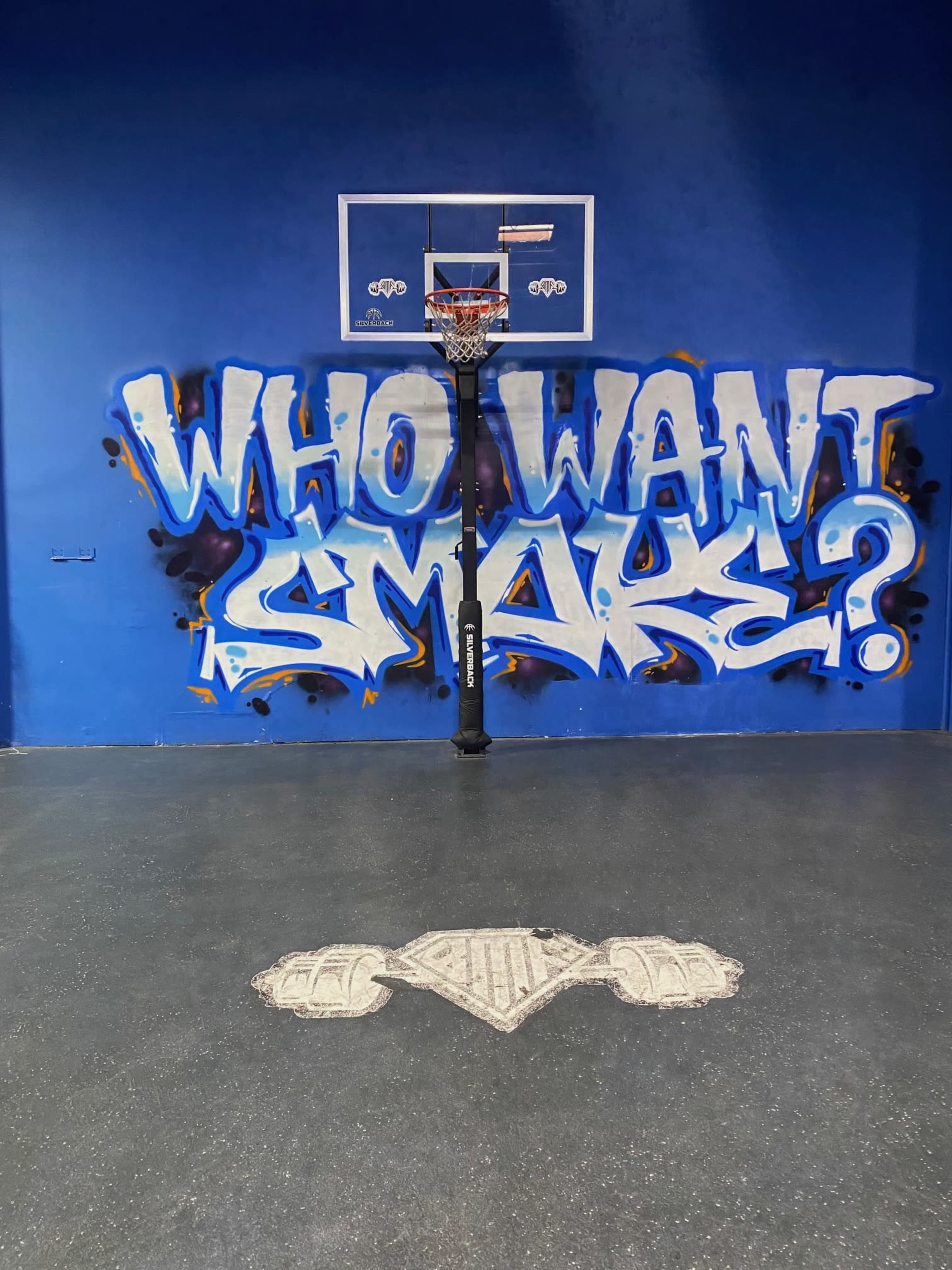 A basketball hoop in front of a blue wall featuring graffiti that reads "WHO WANT SMOKE?"