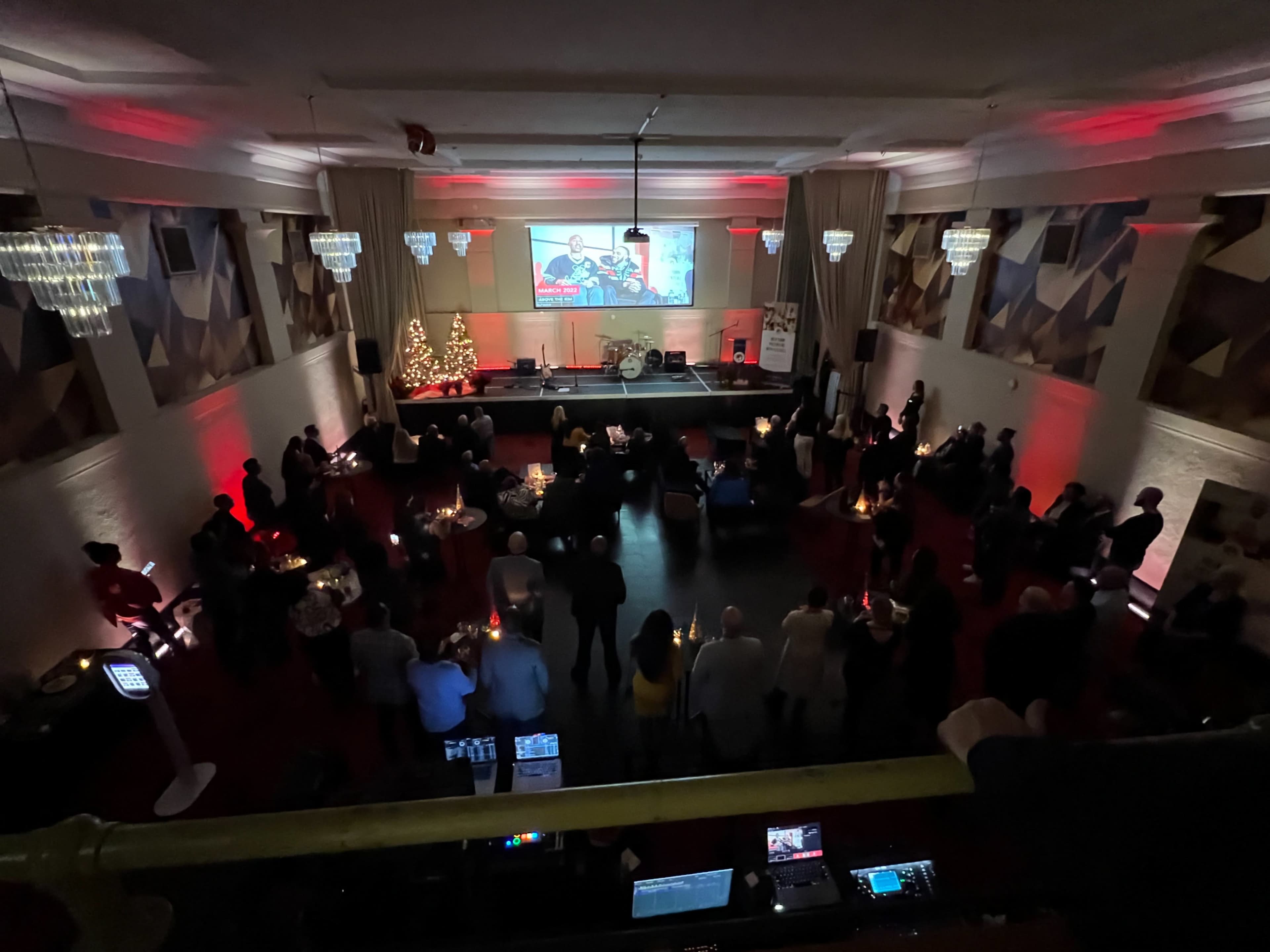 A large group of people gather in a dimly lit venue with a Christmas tree and a screen displaying a festive image.