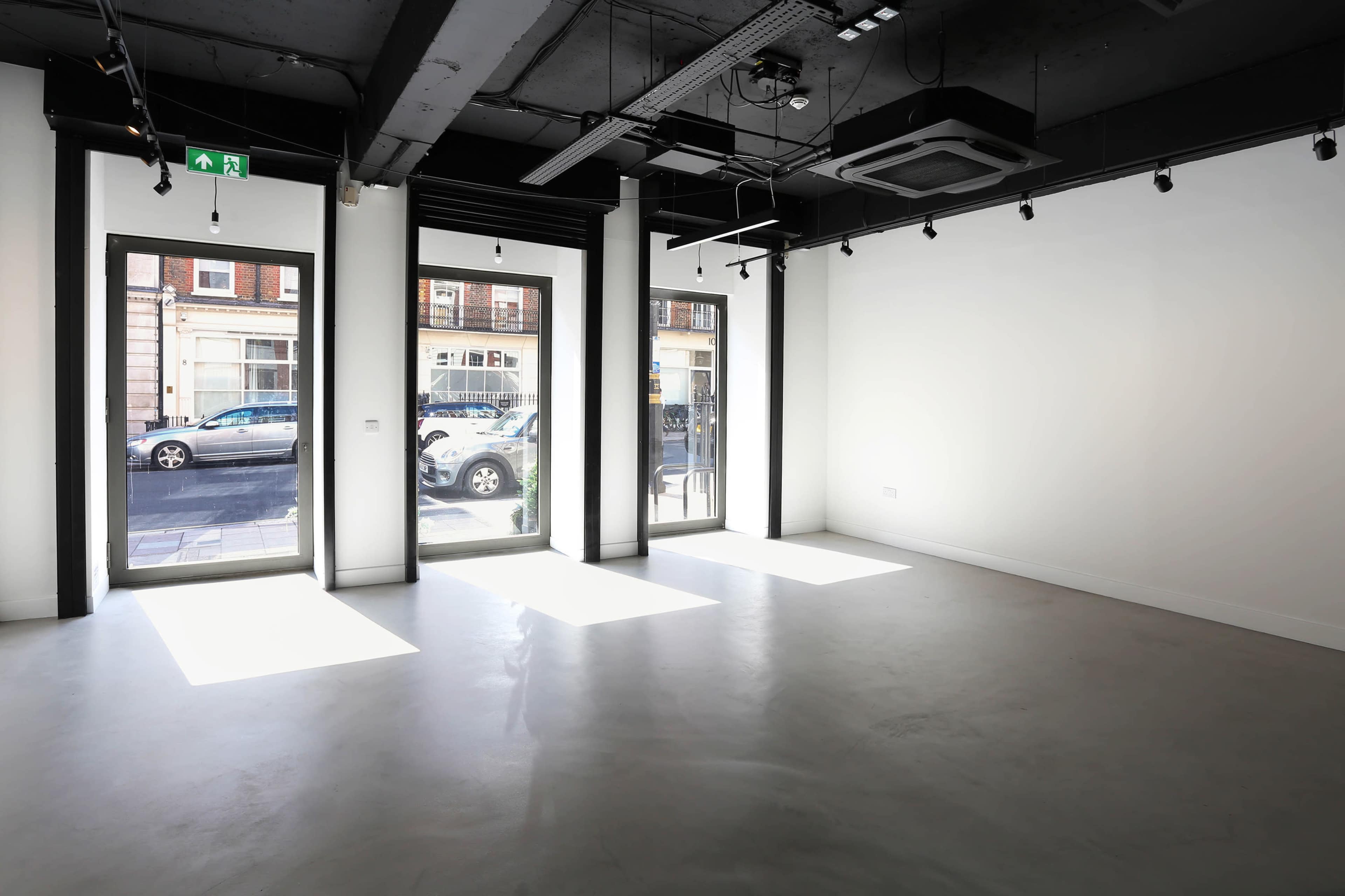The image shows an empty commercial space with large windows allowing natural light to enter, and a clean, minimalist interior featuring a concrete floor.