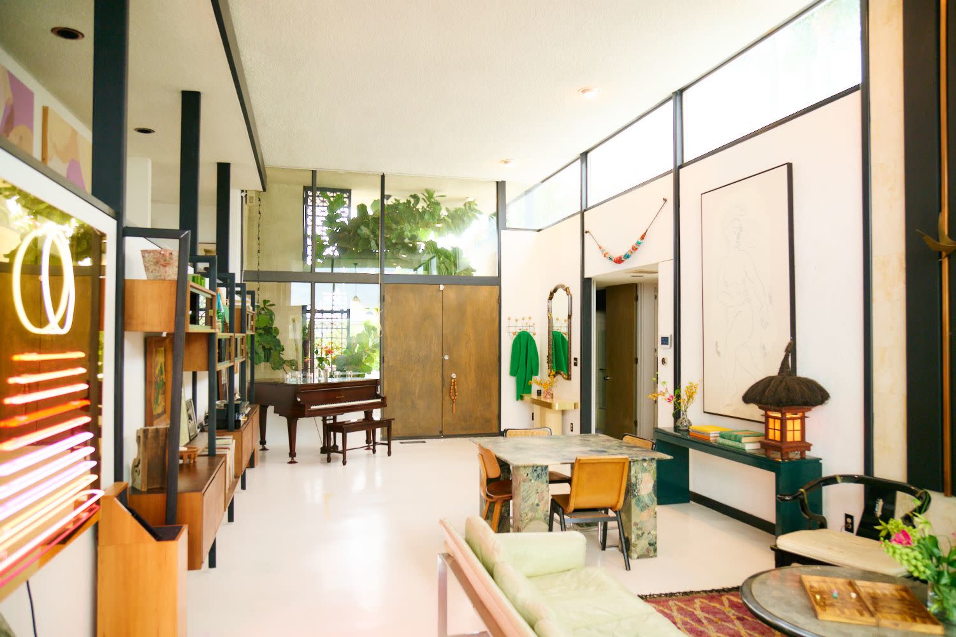 The image features a modern living space with large windows, a piano, and a mix of furniture, including a green sofa and a wooden table, surrounded by indoor plants.