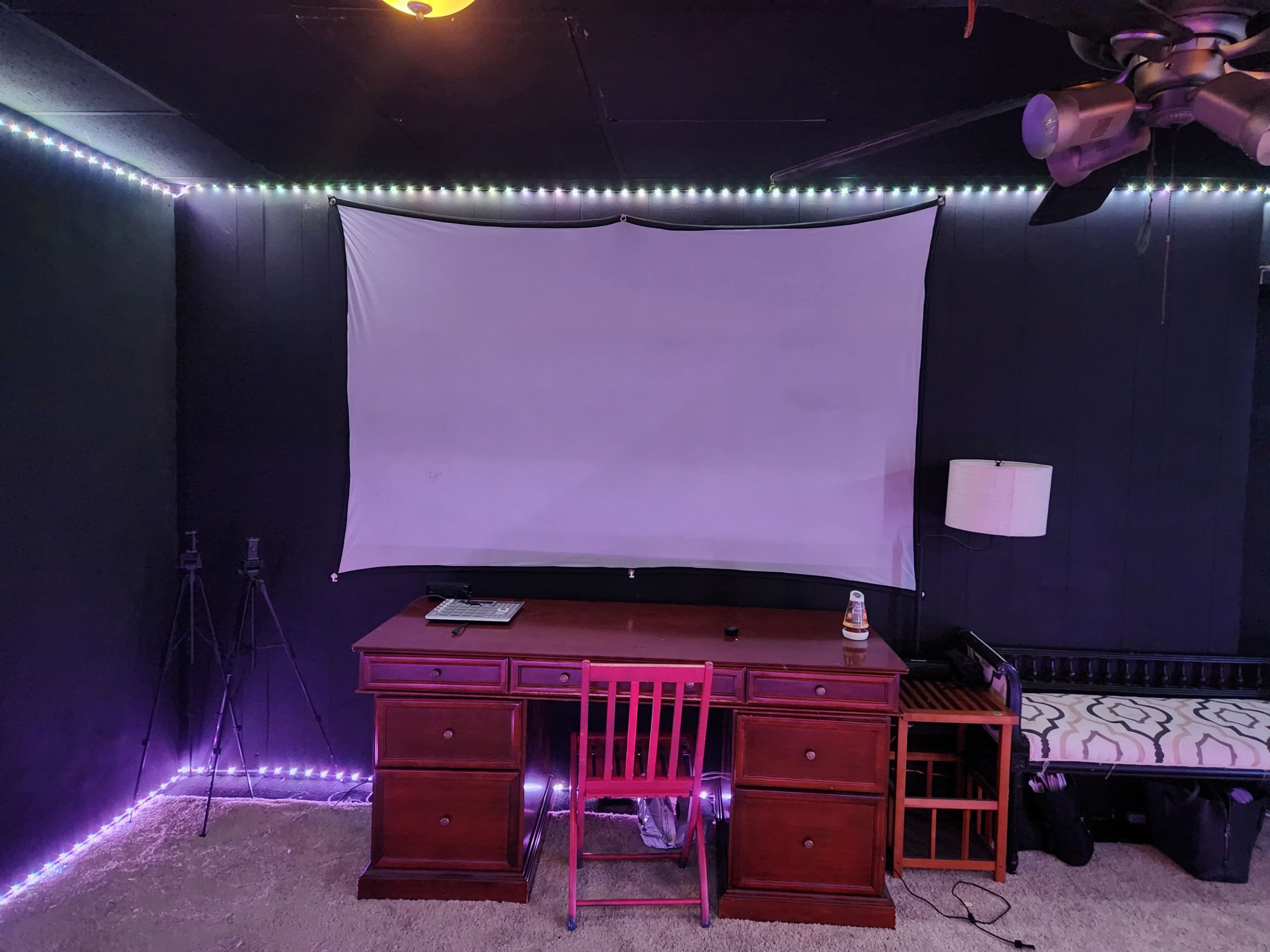 A wooden desk with a pink chair faces a blank projection screen, surrounded by LED lights along the walls in a dimly lit room.