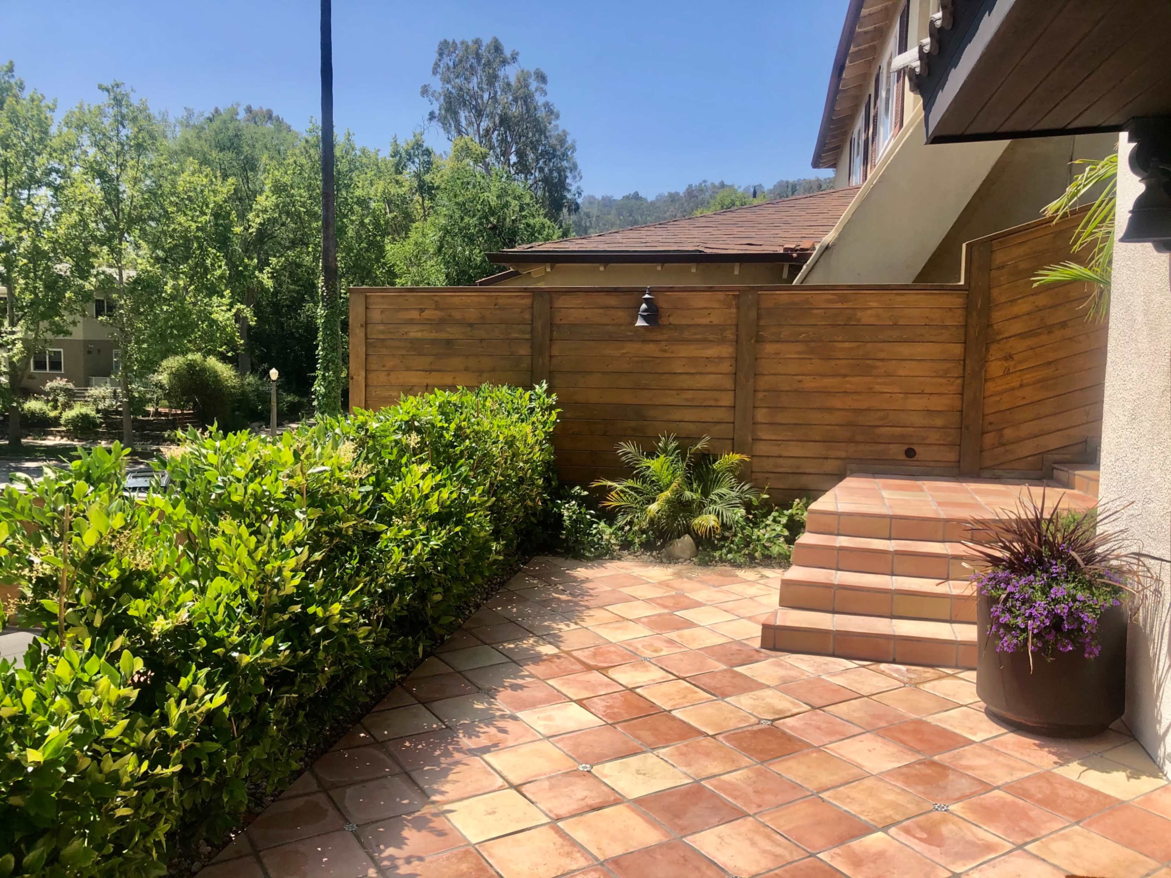 The image shows a brick patio area bordered by a wooden fence and lush greenery, leading to a set of steps.