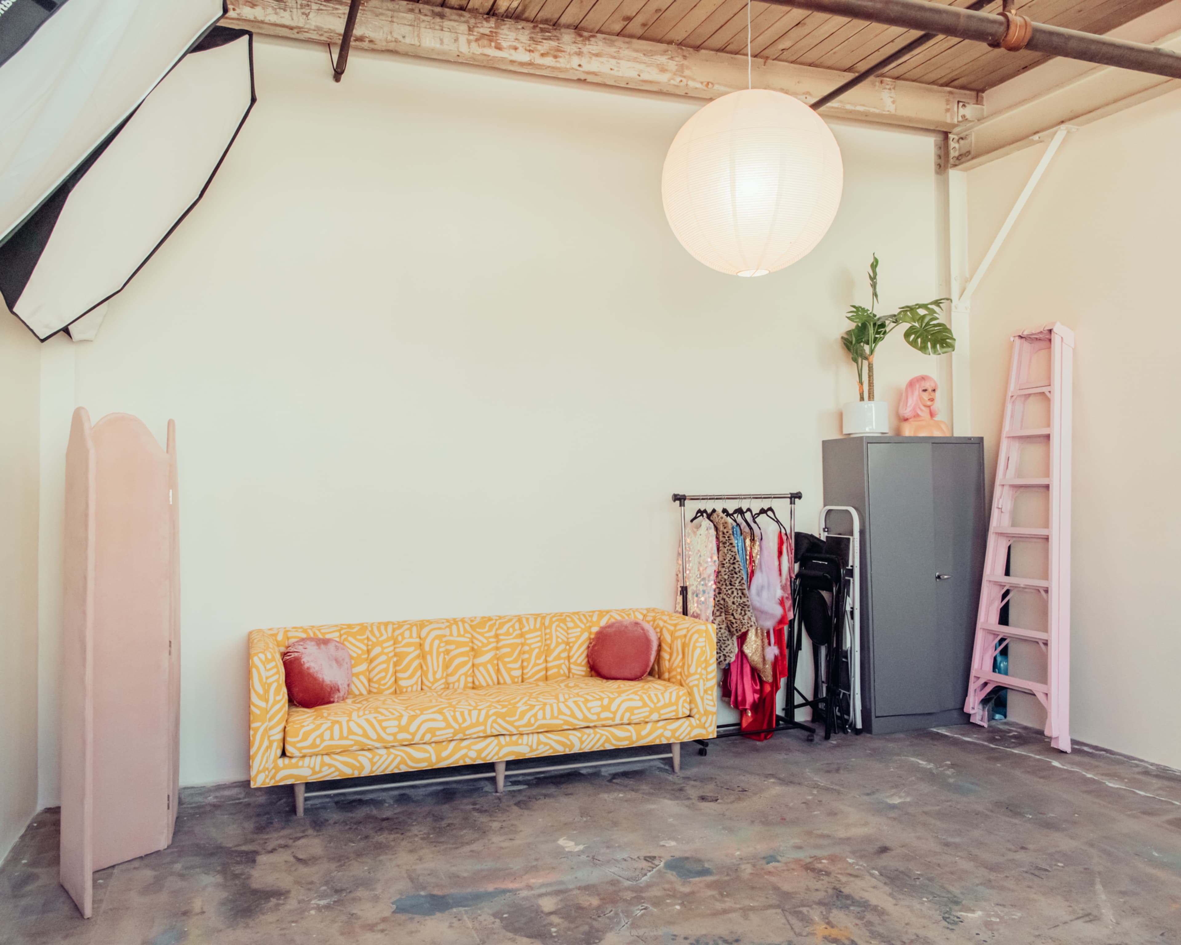 The image shows a minimalist studio space featuring a yellow patterned sofa, a clothing rack, a gray cabinet, a pink ladder, and a round paper lantern hanging from the ceiling.