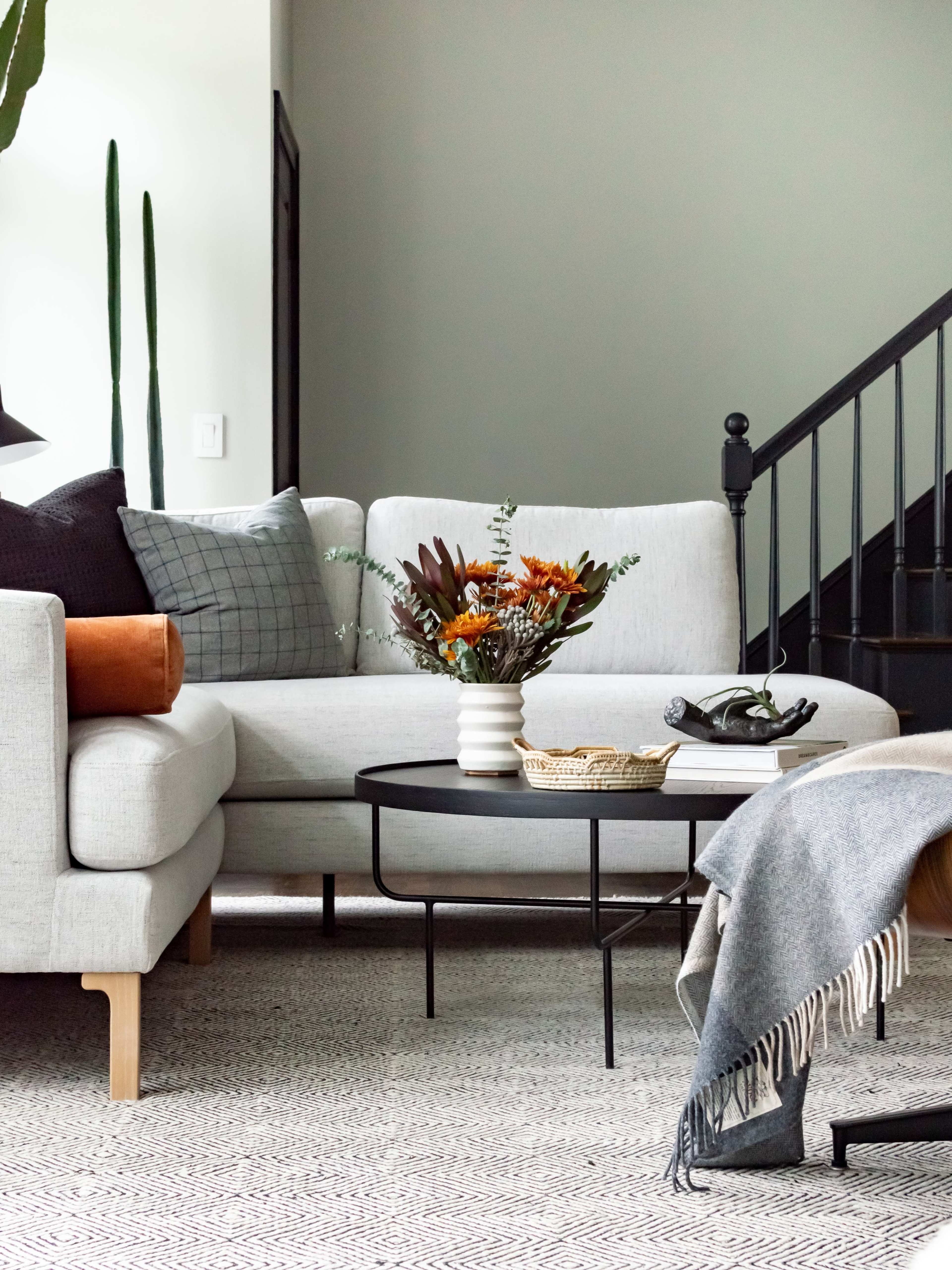 A light gray sectional sofa and a round coffee table are positioned in a modern living room with a staircase in the background.