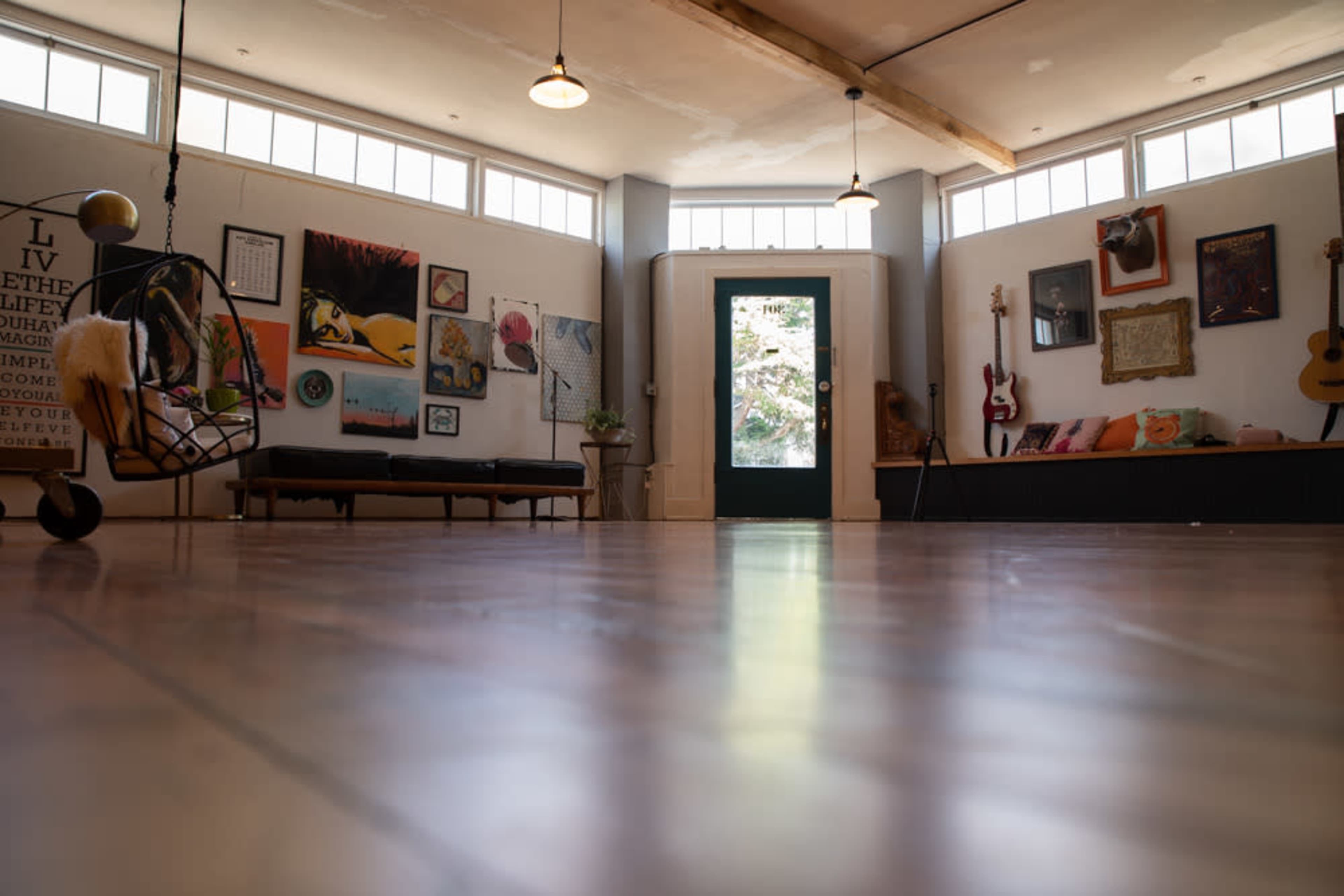 The image shows a bright, spacious interior with wooden flooring, various artworks on the walls, and a green door leading outside.
