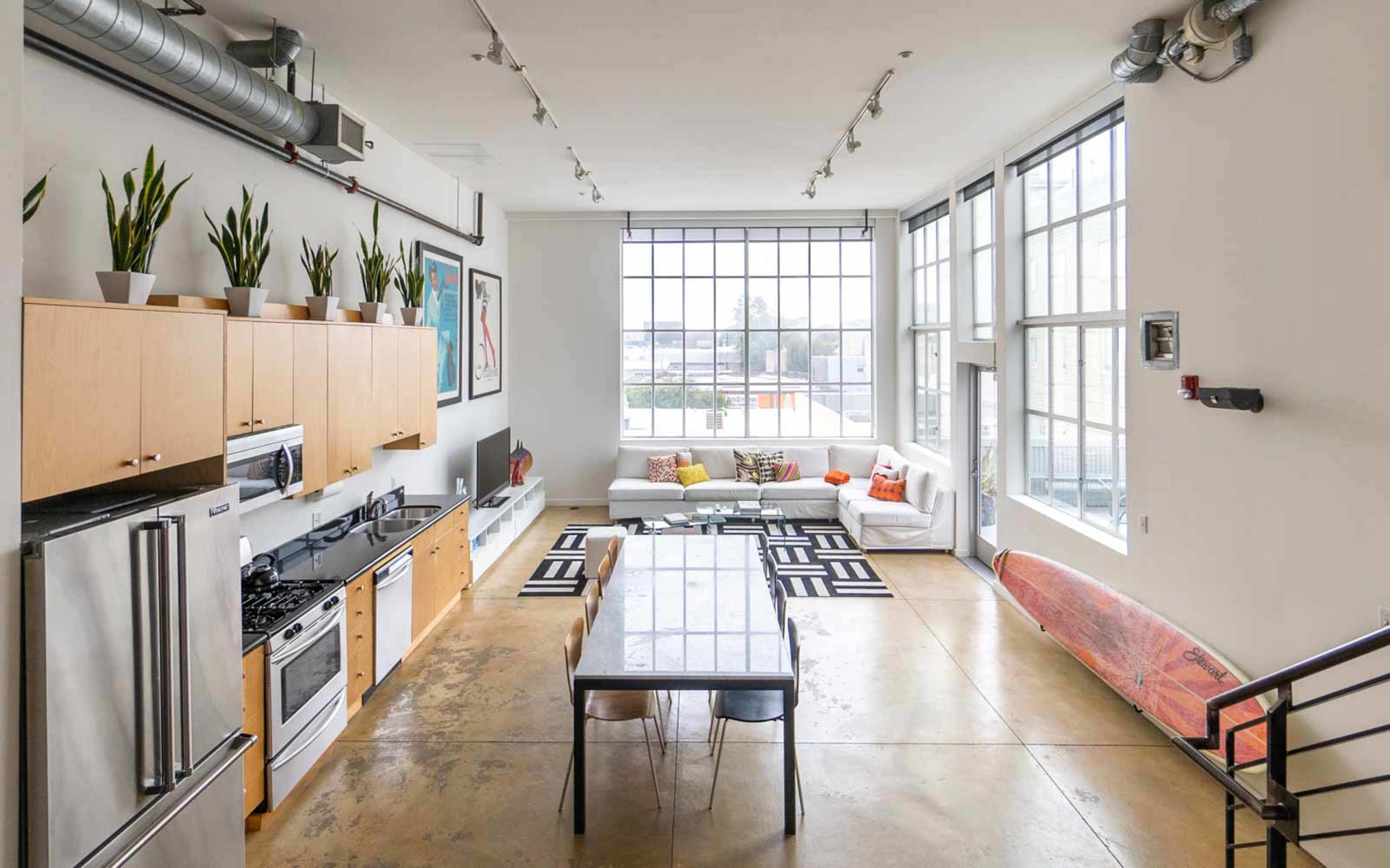 A spacious, modern kitchen and living area with large windows, a dining table, and a surfboard leaning against the wall.