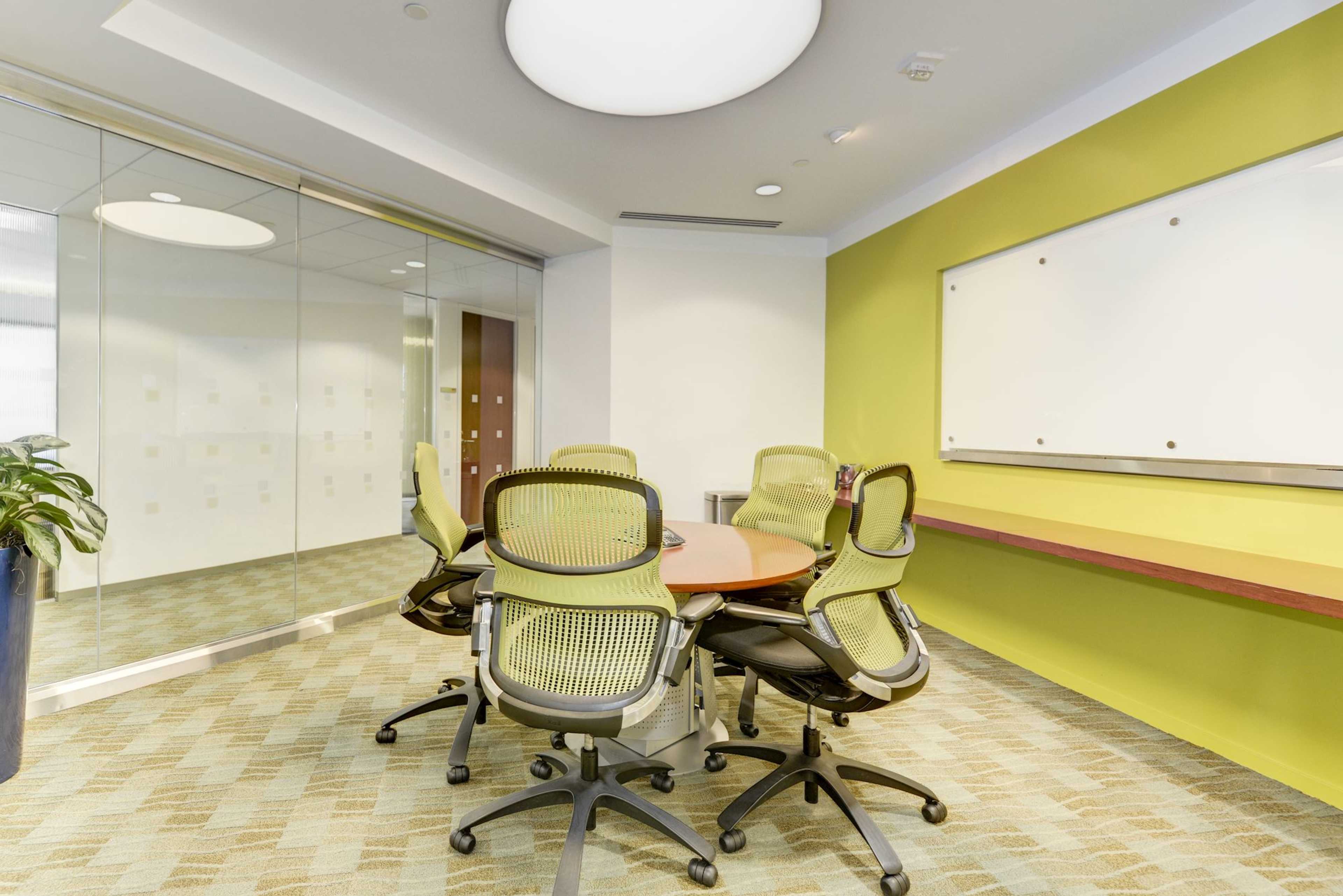 A conference room features a round wooden table surrounded by six green mesh chairs, with a glass wall and a whiteboard on a vibrant green accent wall.