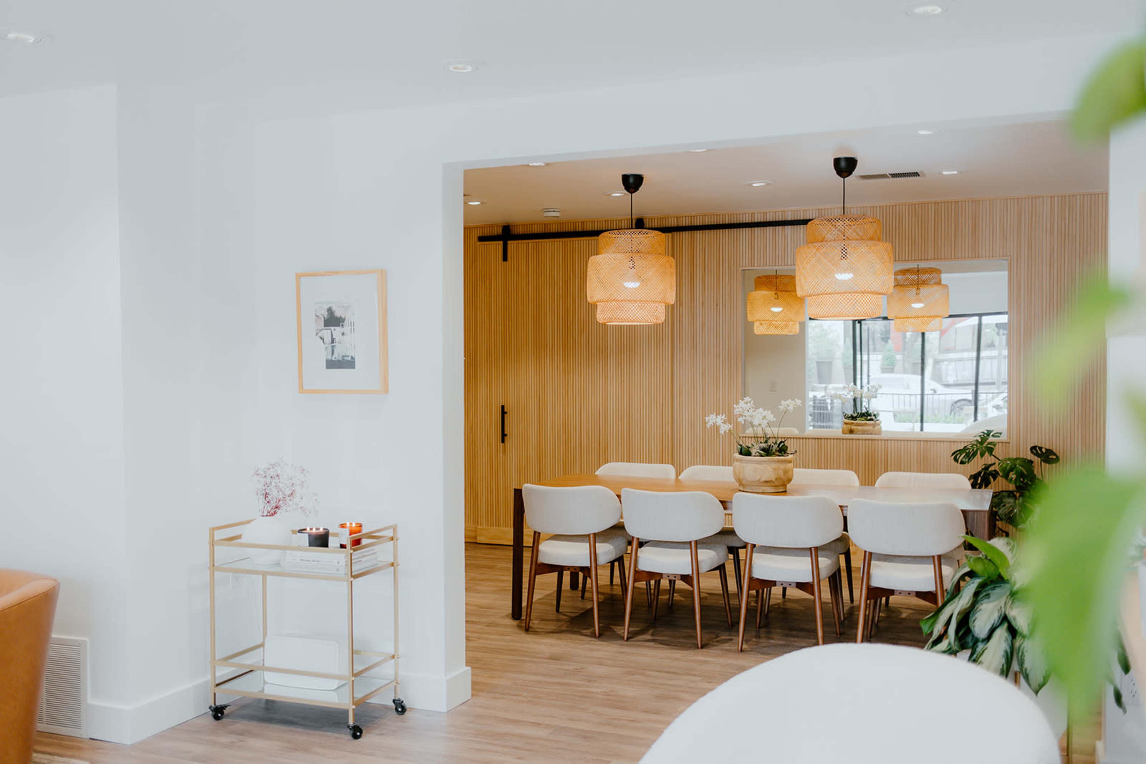 A modern dining area features a wooden table with white chairs, illuminated by pendant lights, and is separated from a living space by a wooden sliding door.