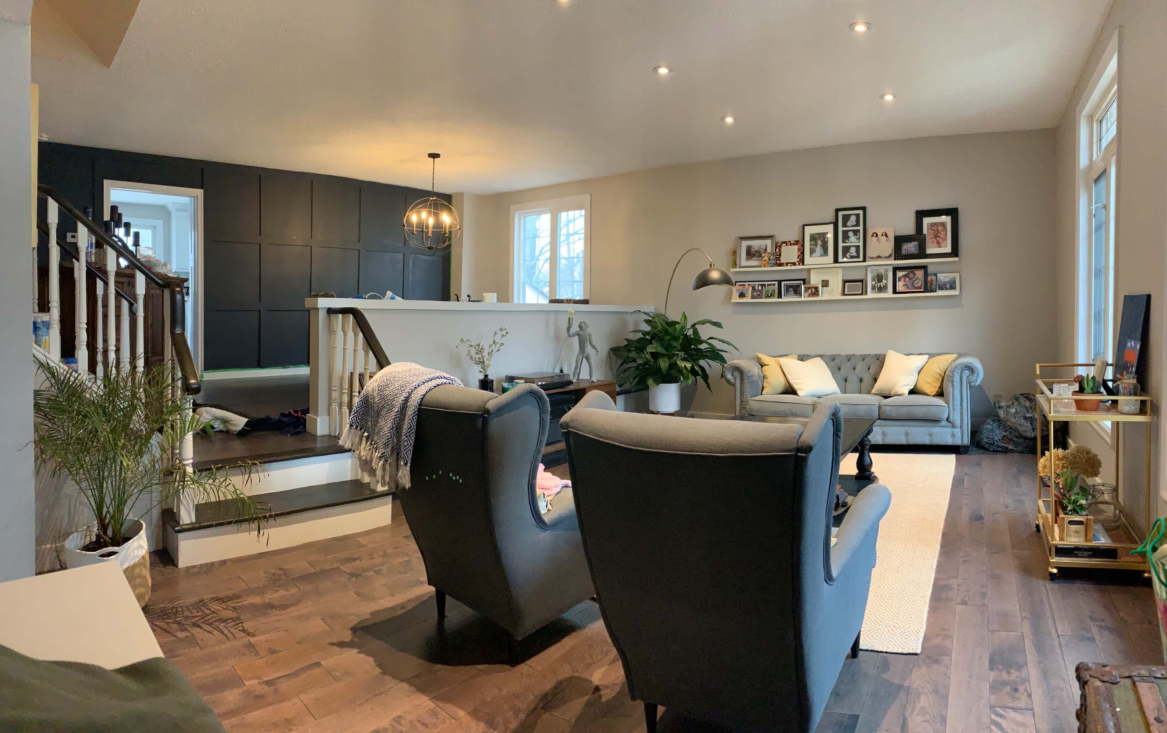 The image shows a modern living room with two armchairs facing a light-colored sofa, a staircase in the background, and a kitchen island visible to the left.