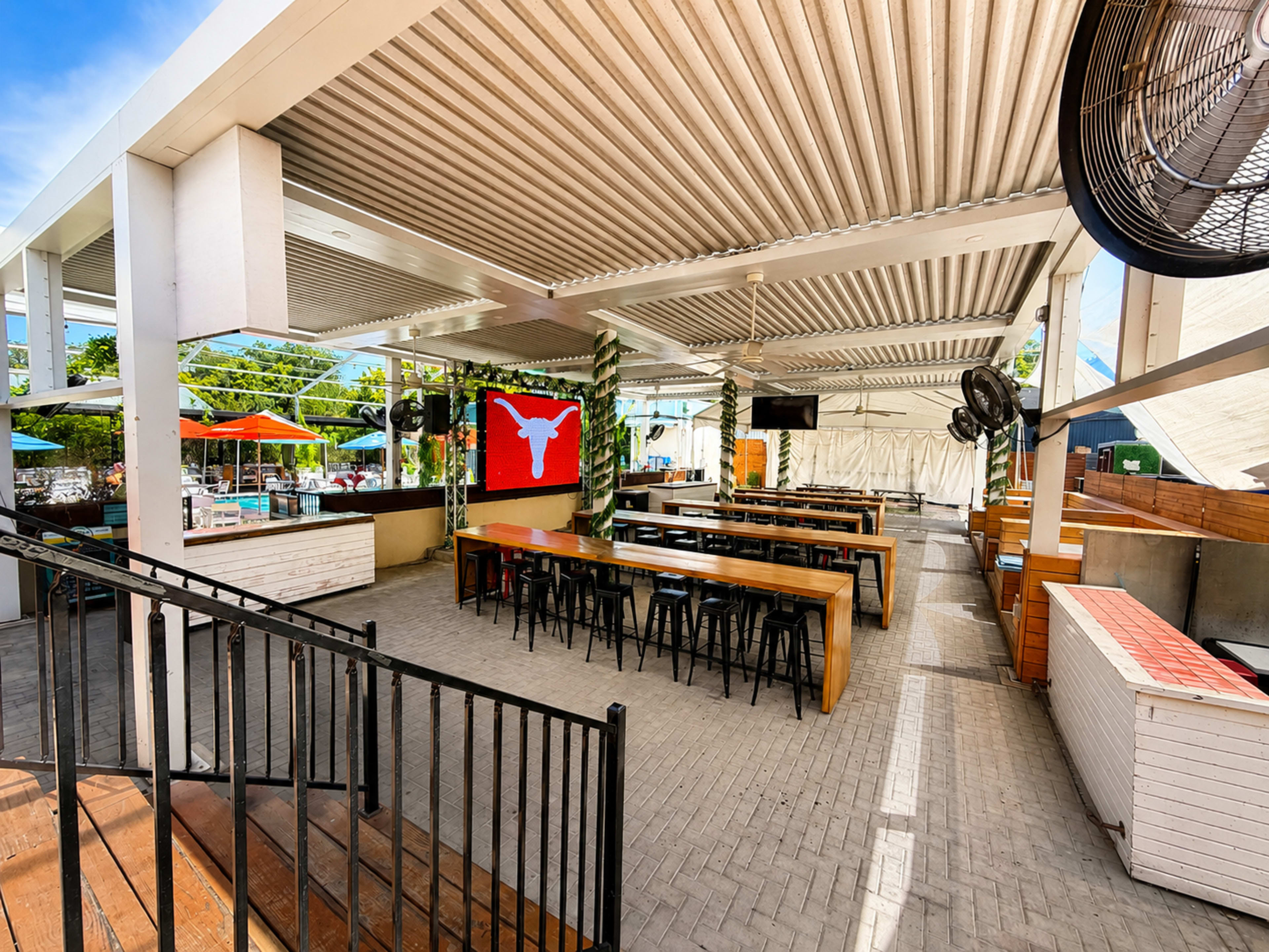 The image shows a spacious outdoor dining area with long wooden tables, black chairs, and a large red flag featuring a white symbol hanging on the wall.