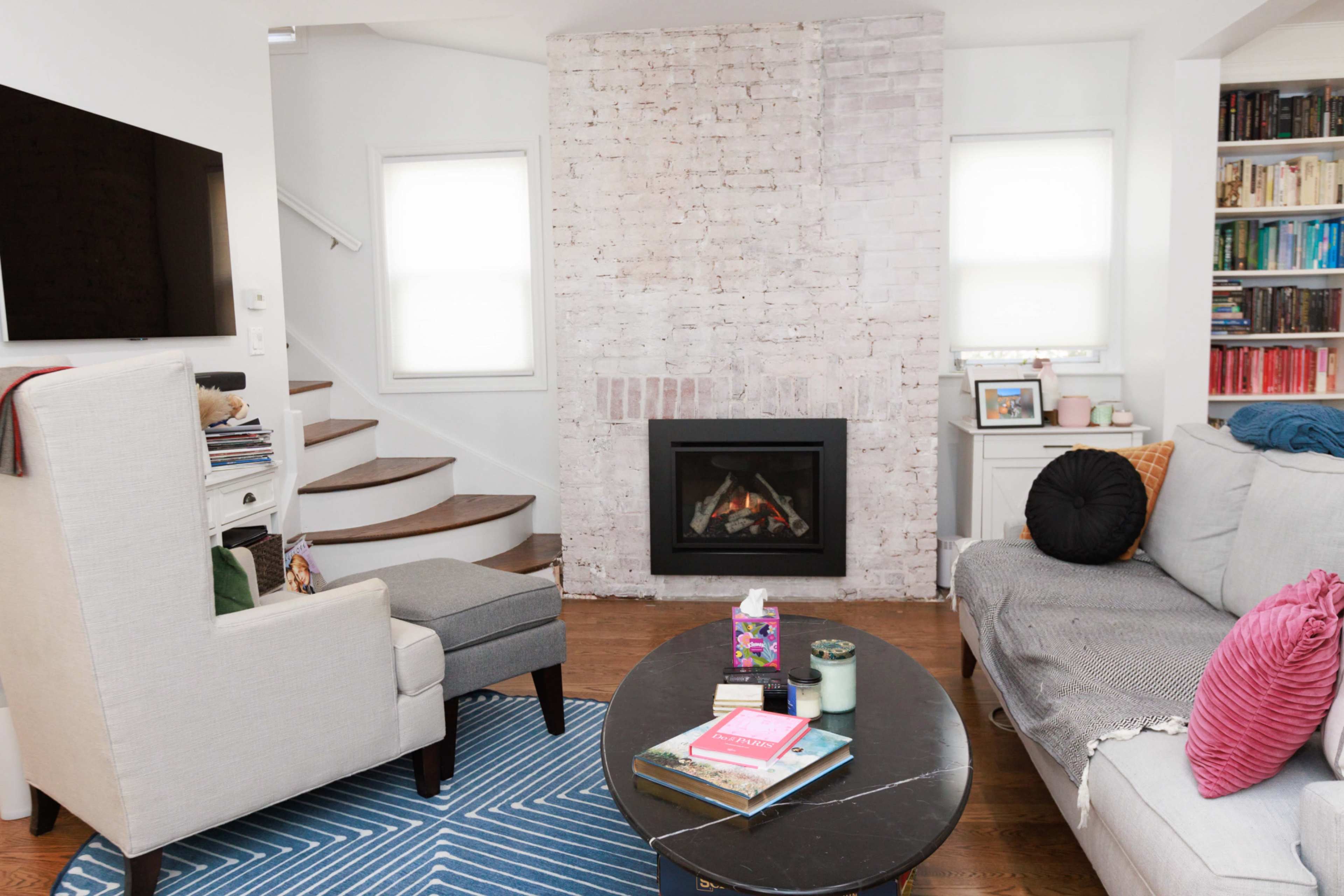 A living room features a sectional sofa, a coffee table with books and candles, a modern fireplace, and a staircase with wooden steps.