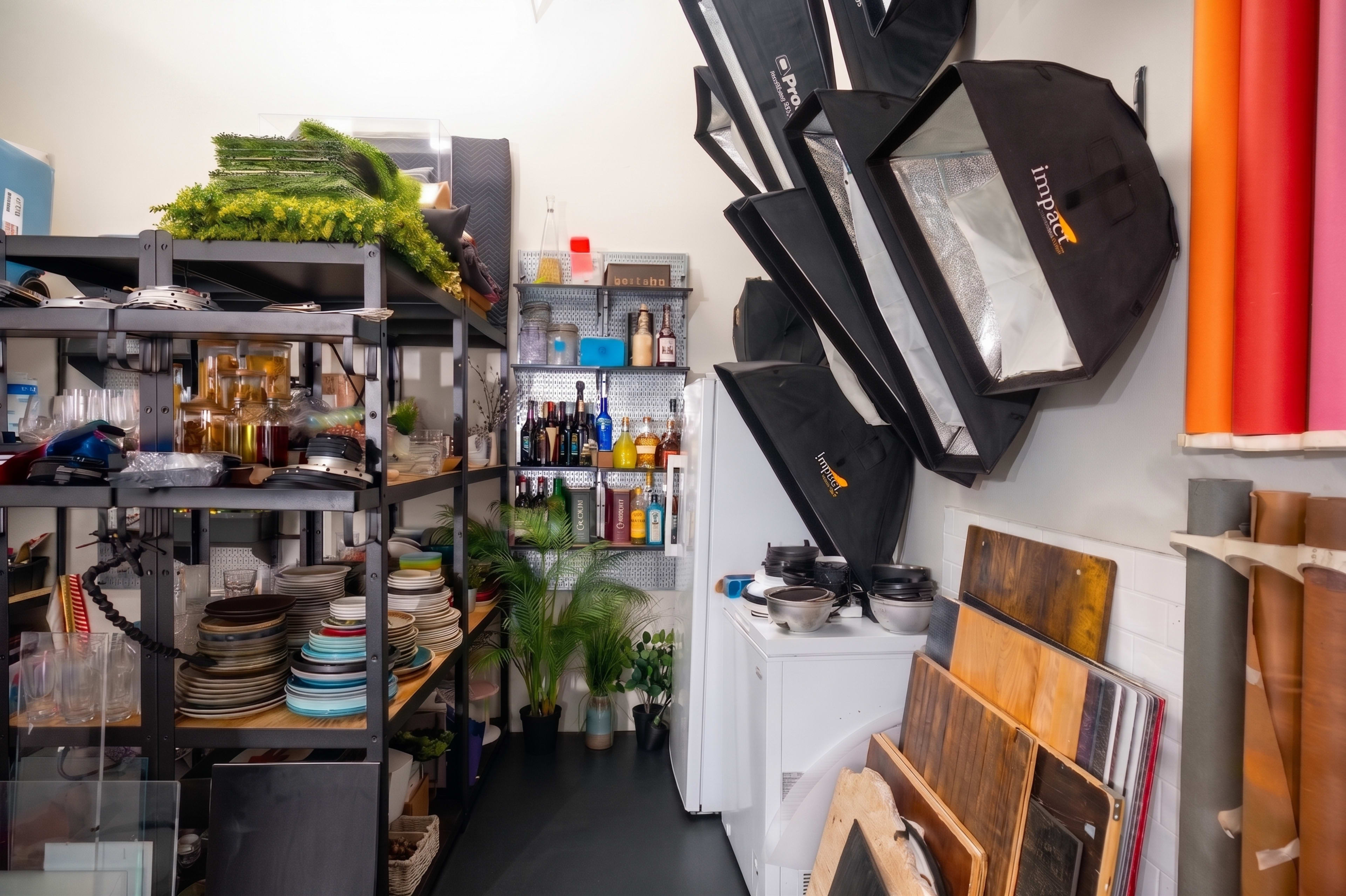 The image shows a storage area filled with shelves of various equipment, dishes, and photography light modifiers, with a washer at the end of the space.