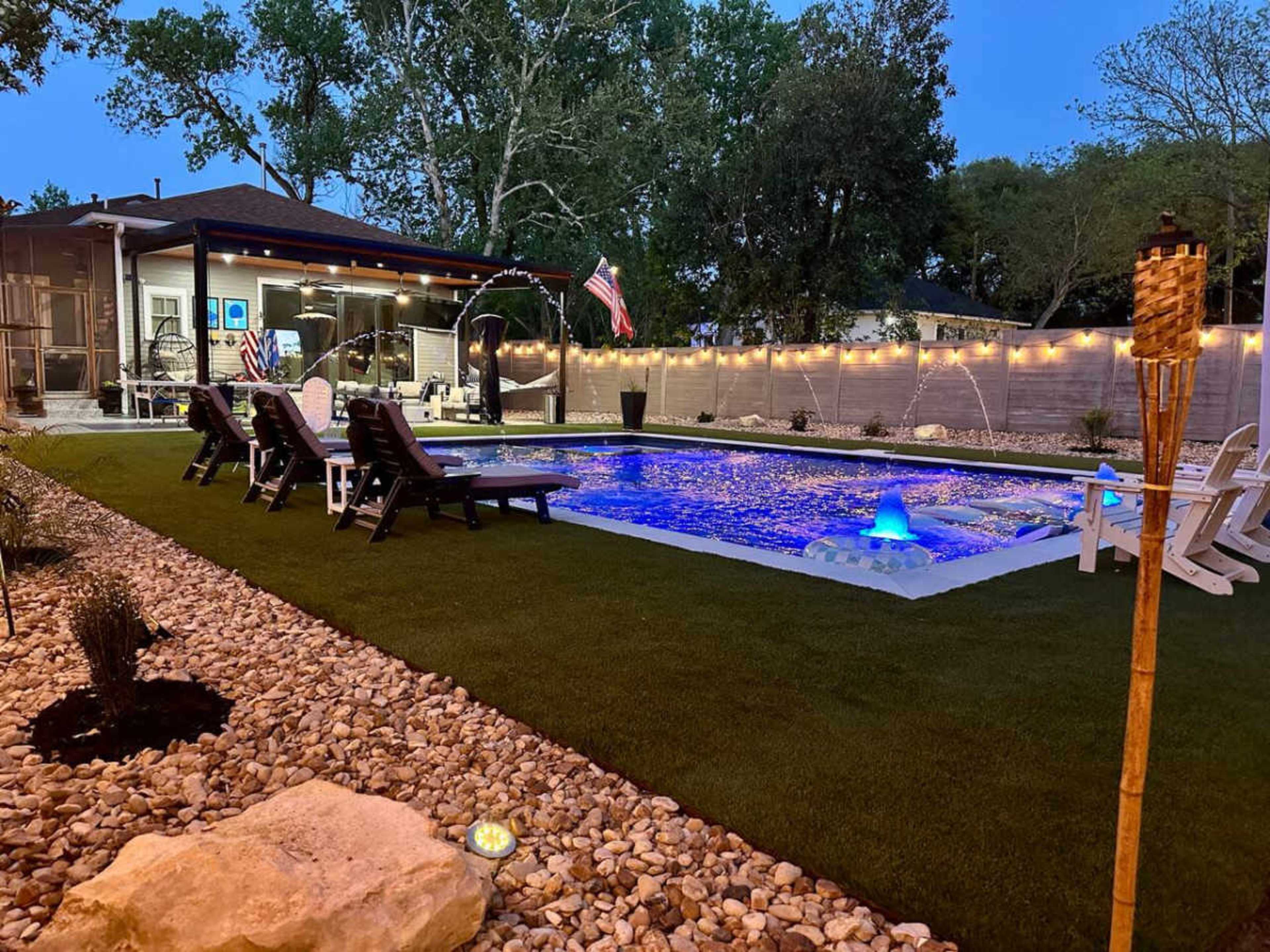A well-lit backyard features a swimming pool with colorful lighting, surrounded by lounge chairs, decorative stones, and a tiki torch.