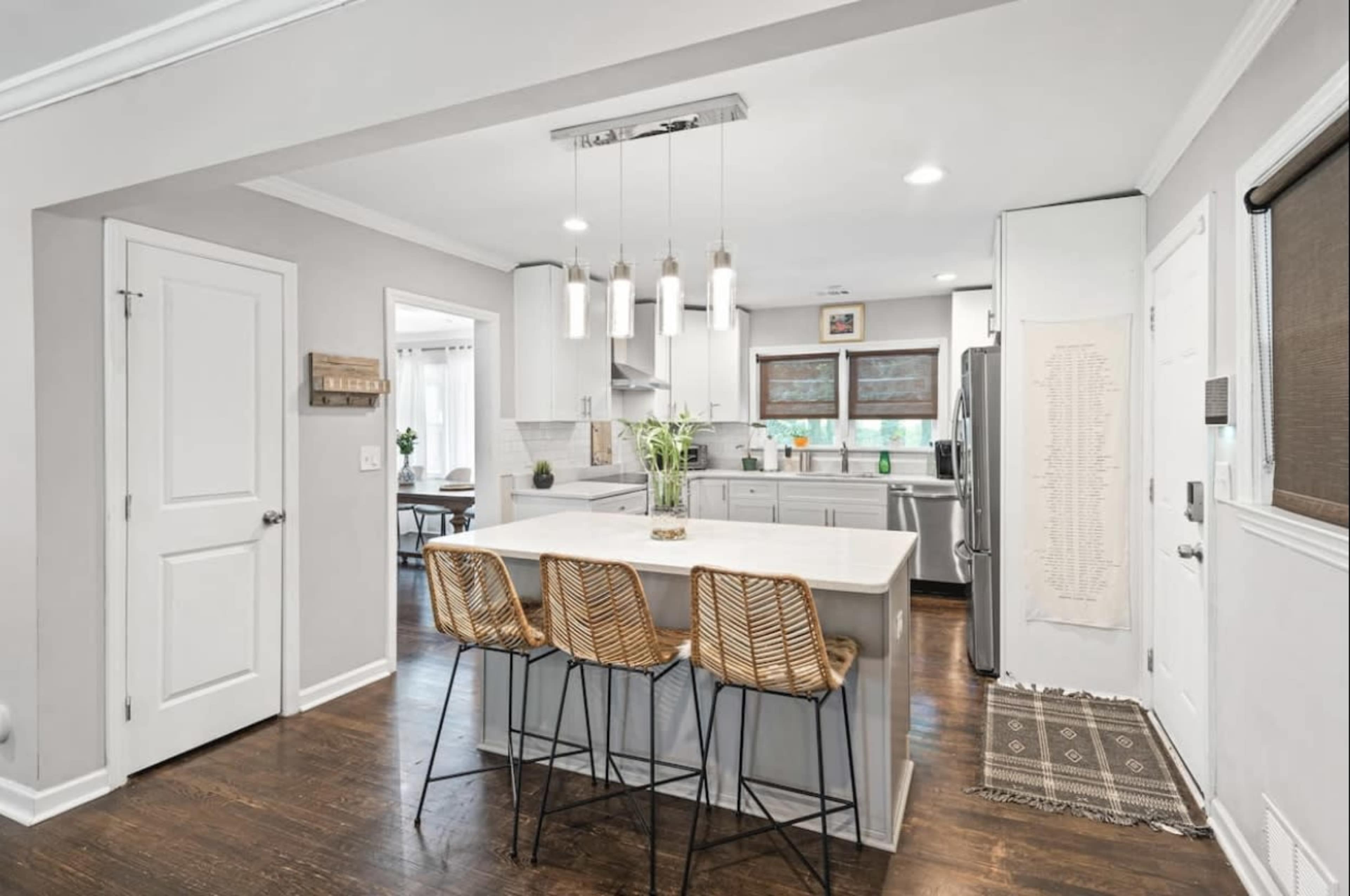 A modern kitchen with a large island and three barstools, featuring white cabinetry, stainless steel appliances, and a dining area in the background.