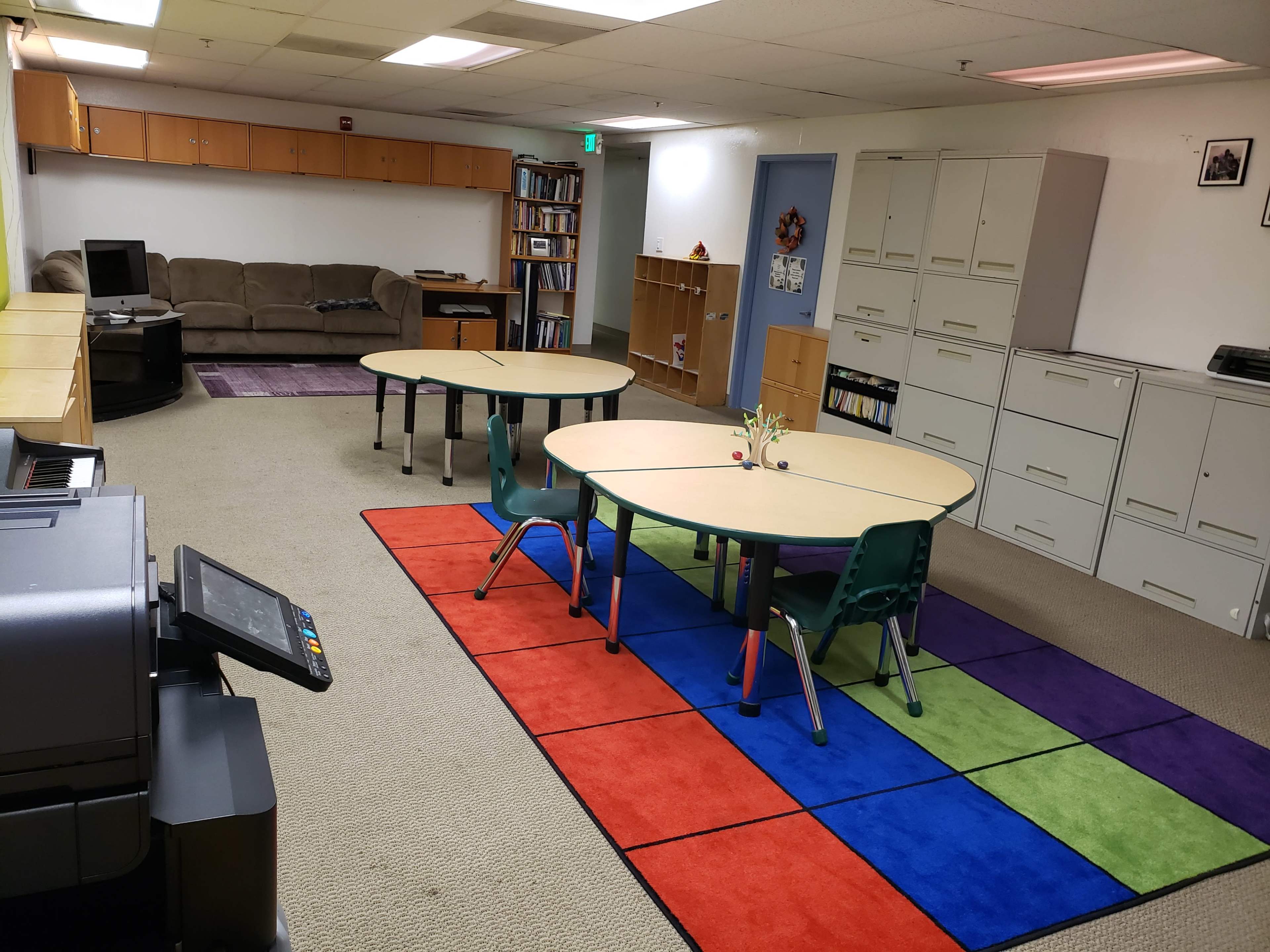 A classroom with two circular tables and chairs on a colorful carpet, alongside storage cabinets and a couch in the background.