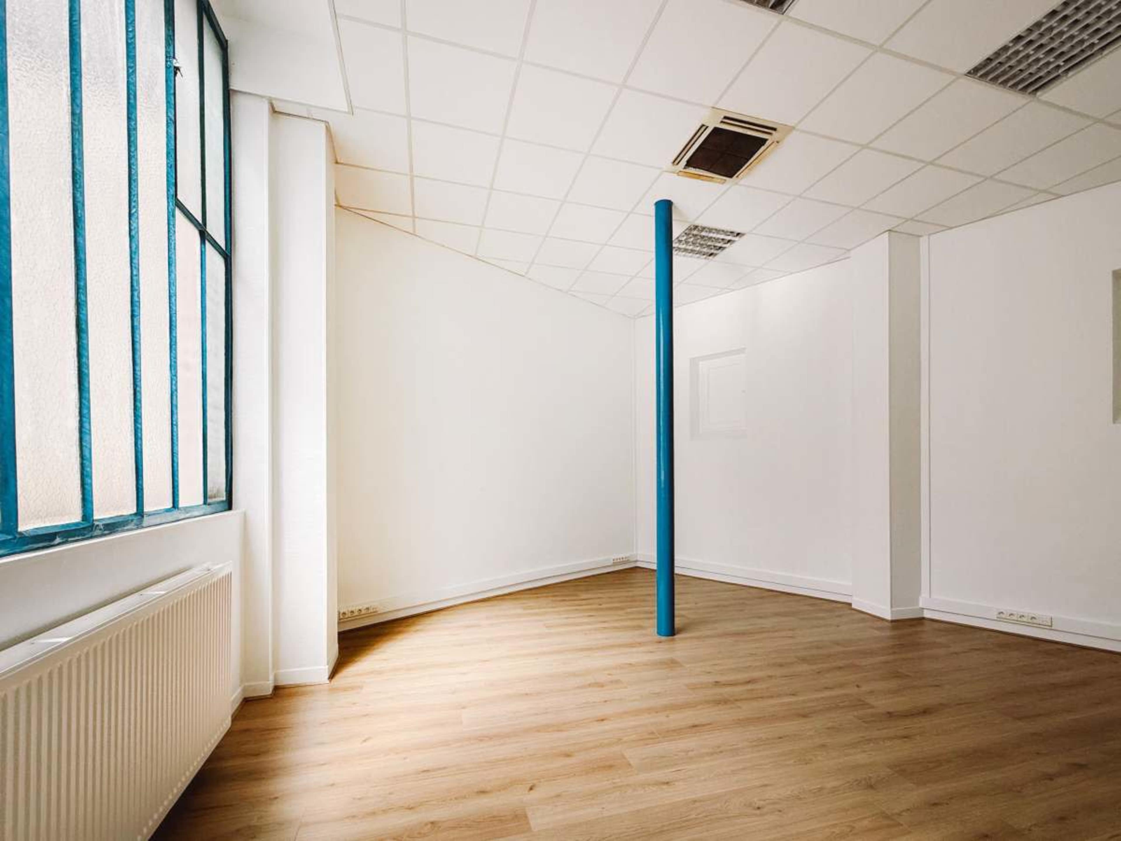 The image shows an empty room with white walls, a wooden floor, a blue vertical pole in the center, and a large window on one side.
