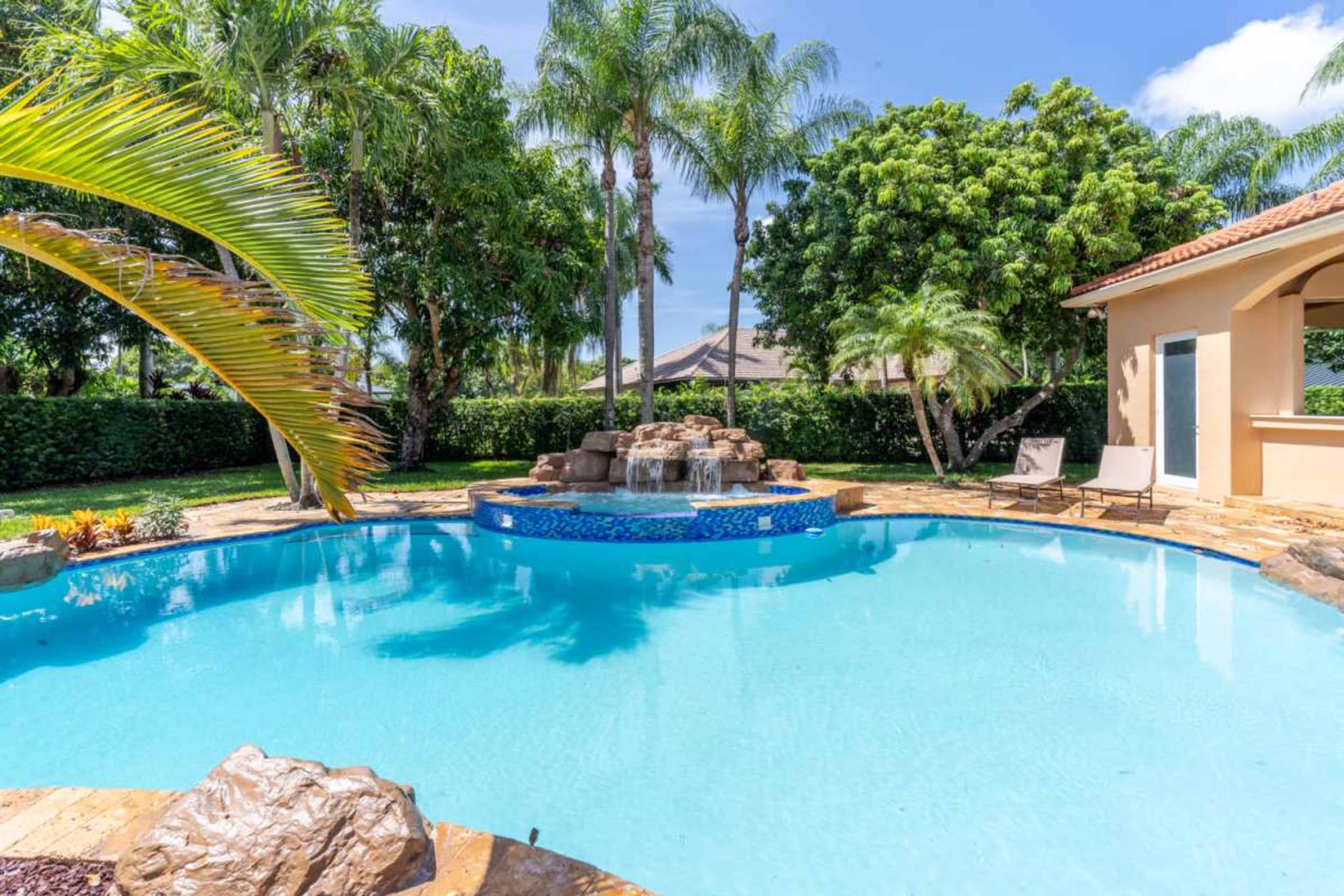 The image shows a residential backyard featuring a swimming pool with a rock waterfall, surrounded by palm trees and landscaped greenery.