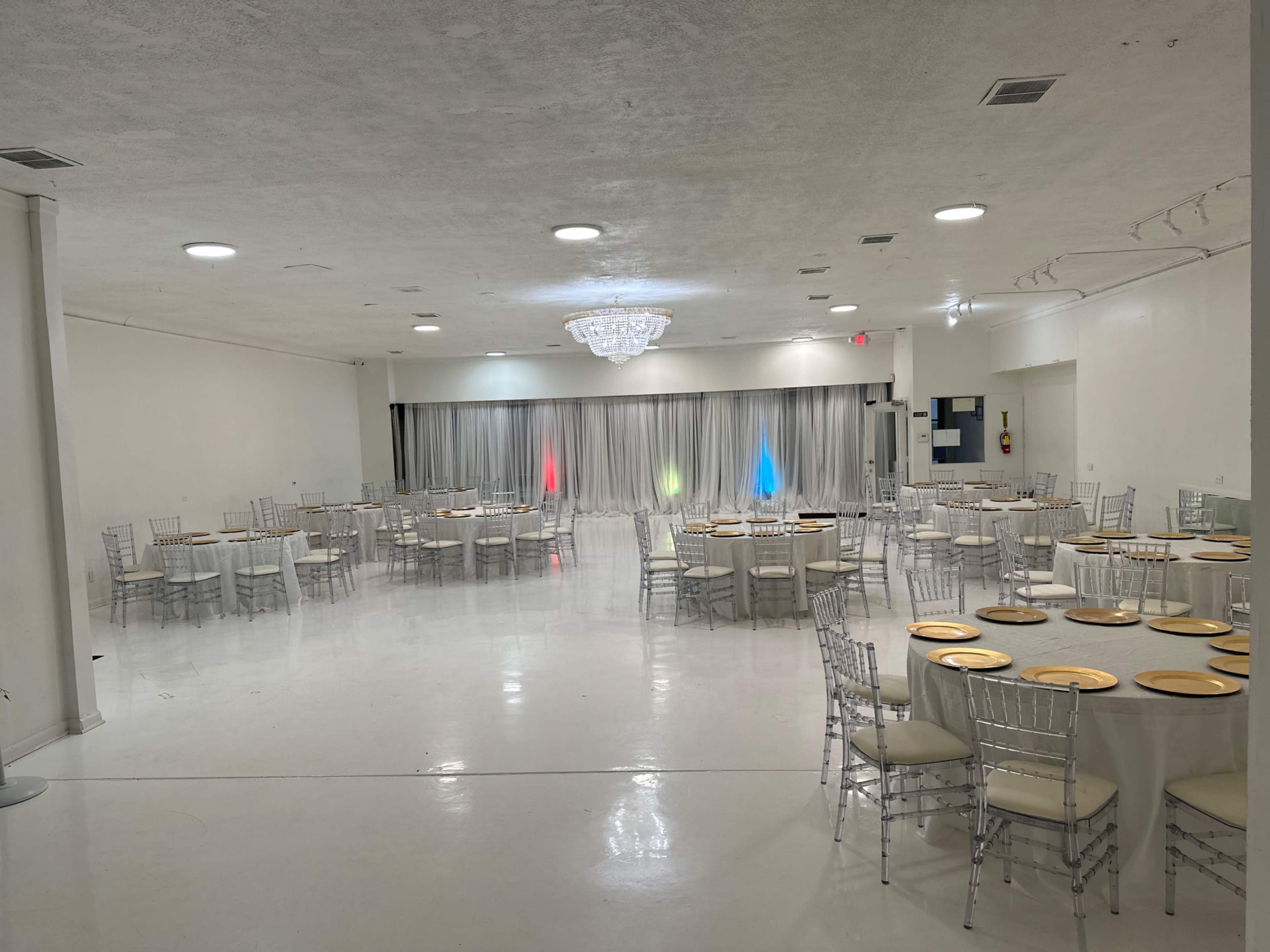 A spacious event hall features round tables with white tablecloths, set with gold chargers, and a backdrop of colored lights behind a curtain.