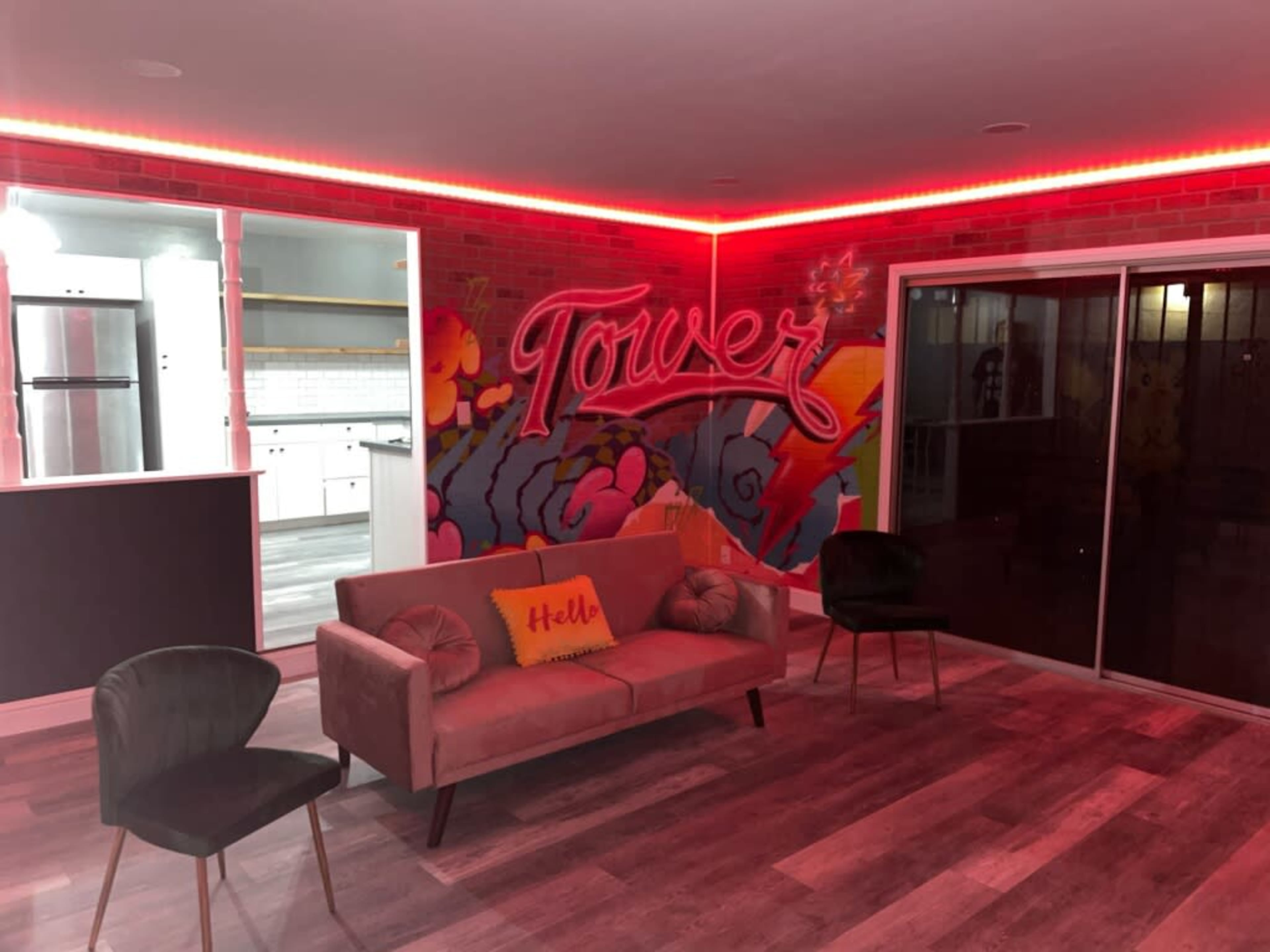 A modern living space with a pink sofa, two chairs, and a colorful mural on a brick wall, illuminated by red LED lights.