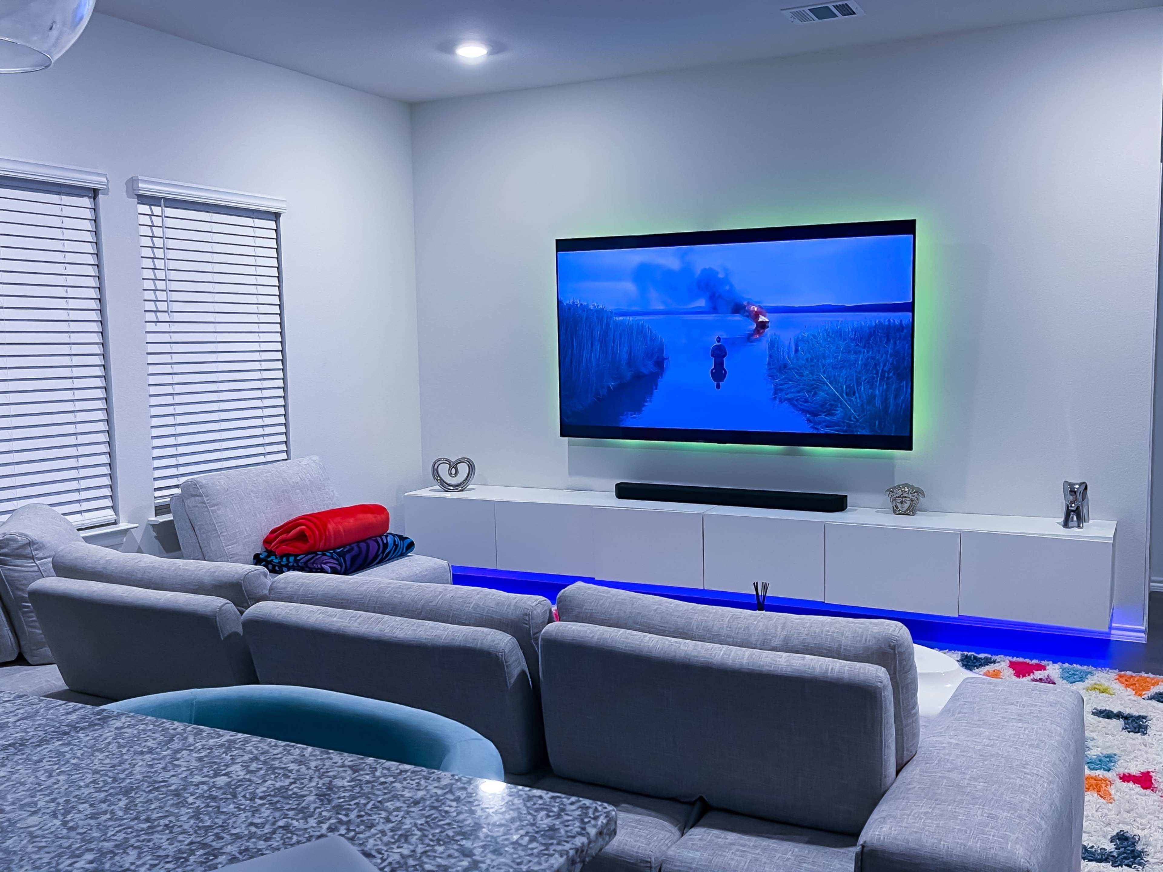 A modern living room features a large television displaying a scene from a movie, with a grey sectional sofa and a lighted entertainment unit underneath.