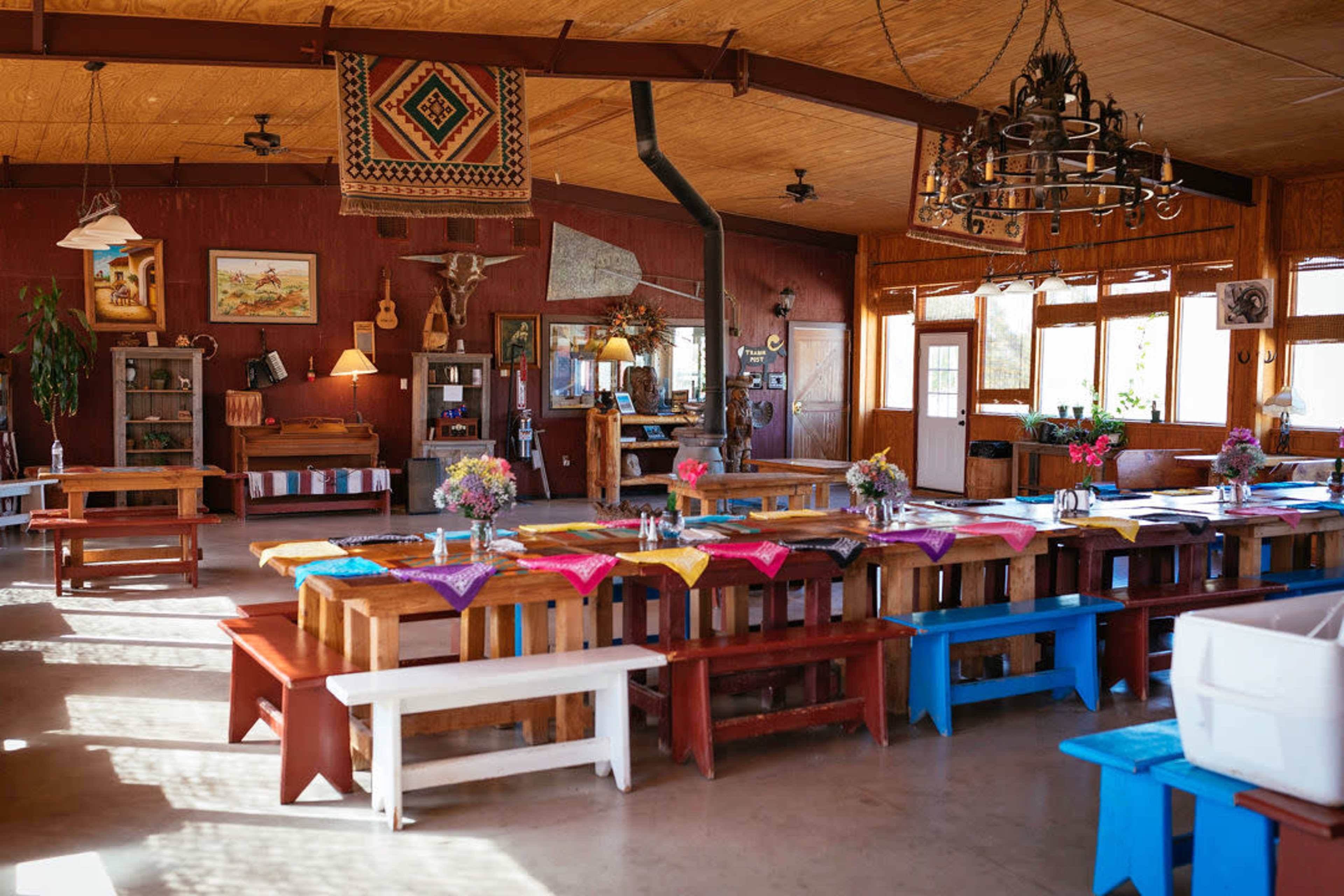 The interior of a rustic dining hall features multiple long wooden tables adorned with colorful table runners and centerpieces, illuminated by natural light from large windows.