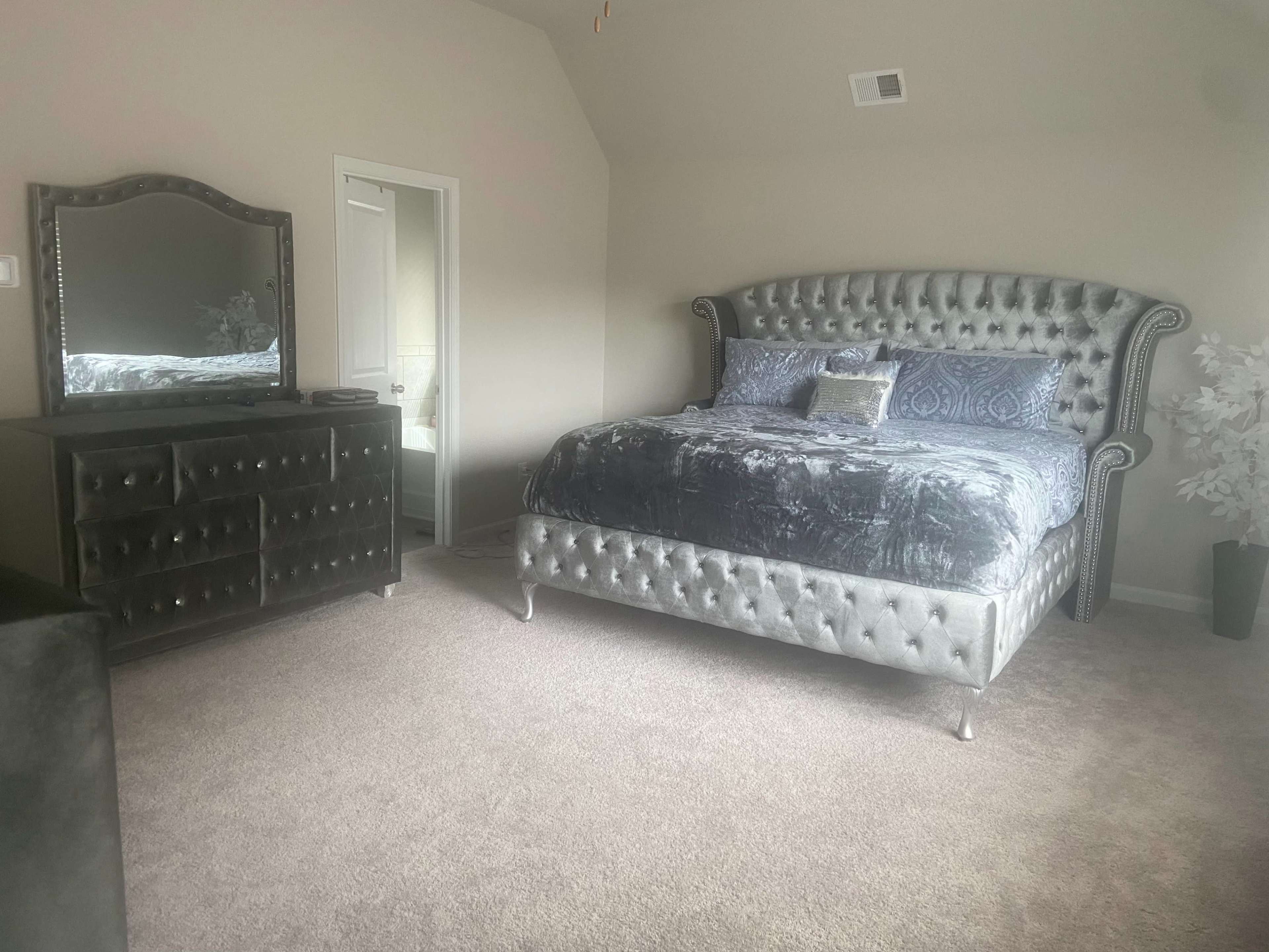 A bedroom with a tufted gray bed, a matching dresser, and a mirror, all set against light-colored walls and carpet.