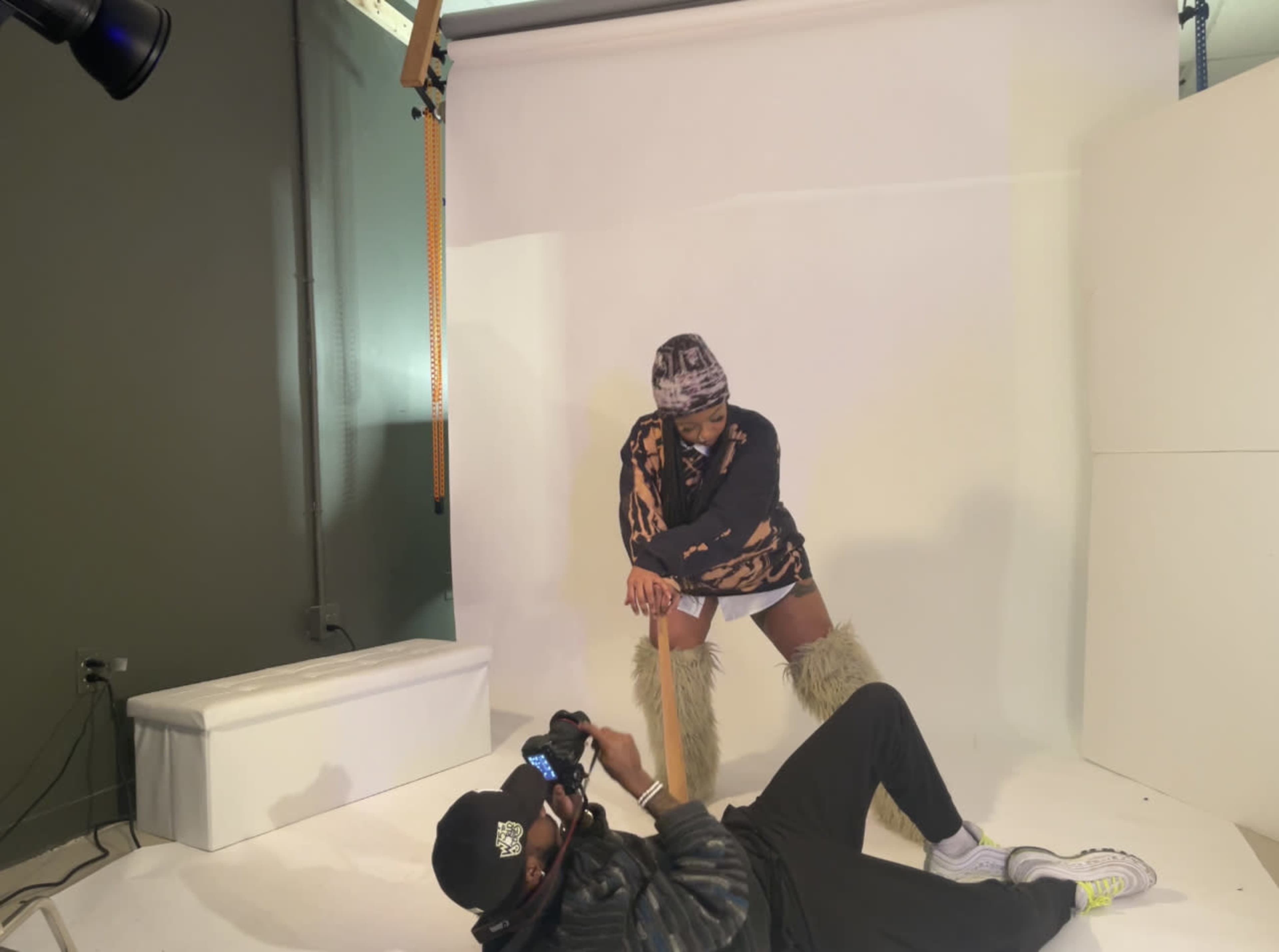 A person poses on a white backdrop while another person lies on the ground taking photos.