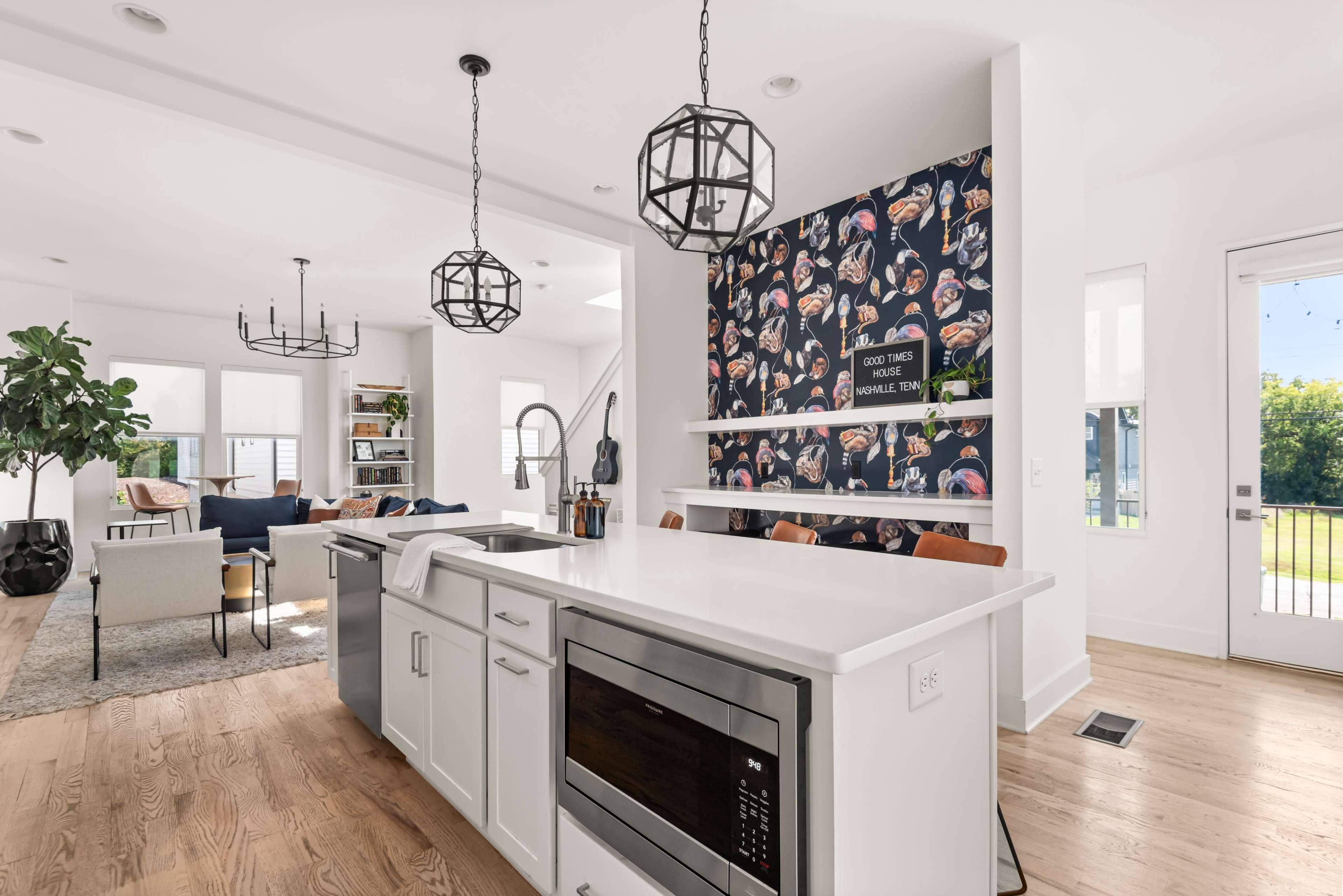The image shows a modern kitchen with a white countertop, dark cabinetry, colorful wallpaper featuring food illustrations, and a view of an open living area.