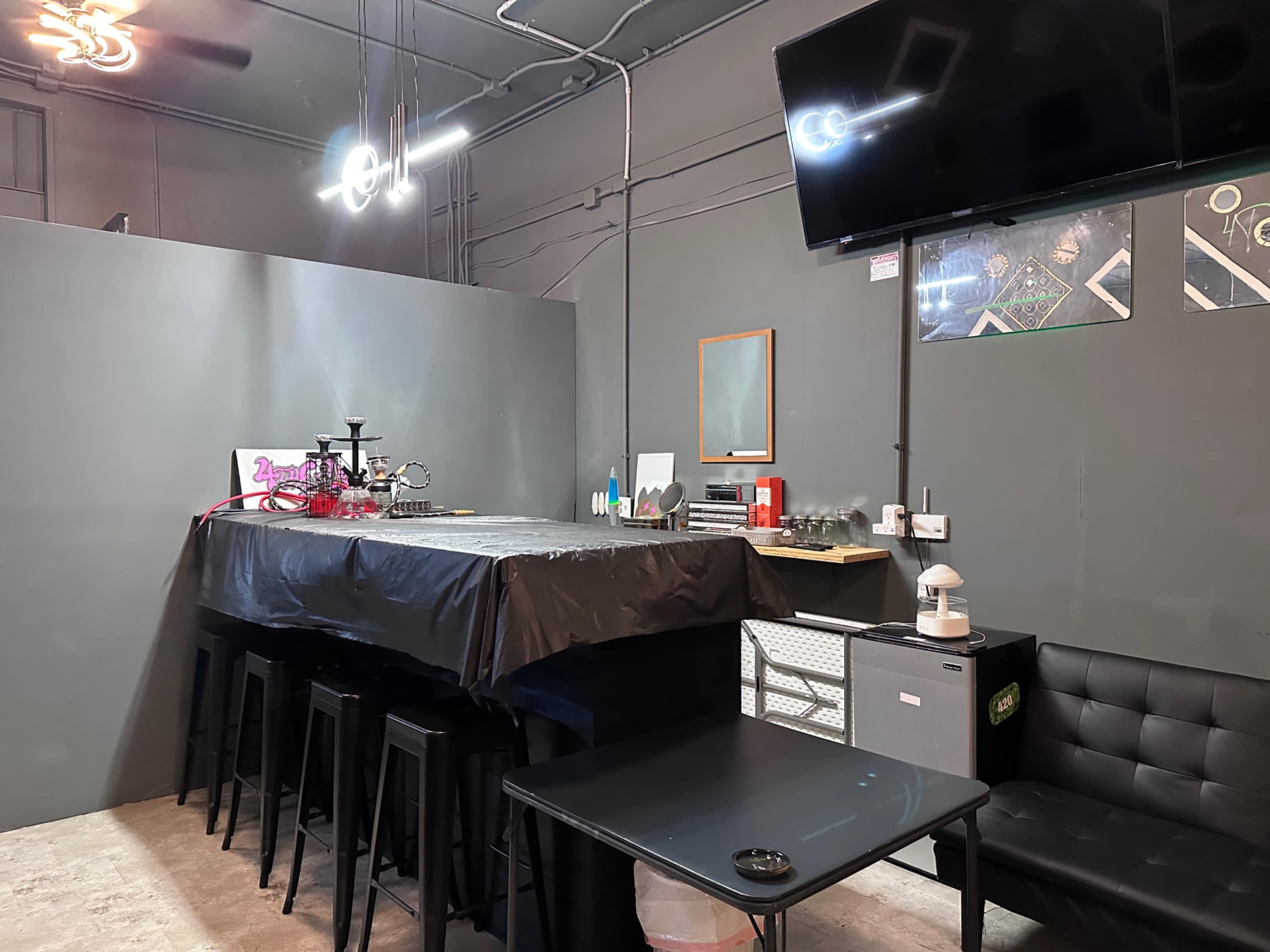 The image shows a modern lounge area with a long black table, chairs, a hookah setup, and a television mounted on the wall.