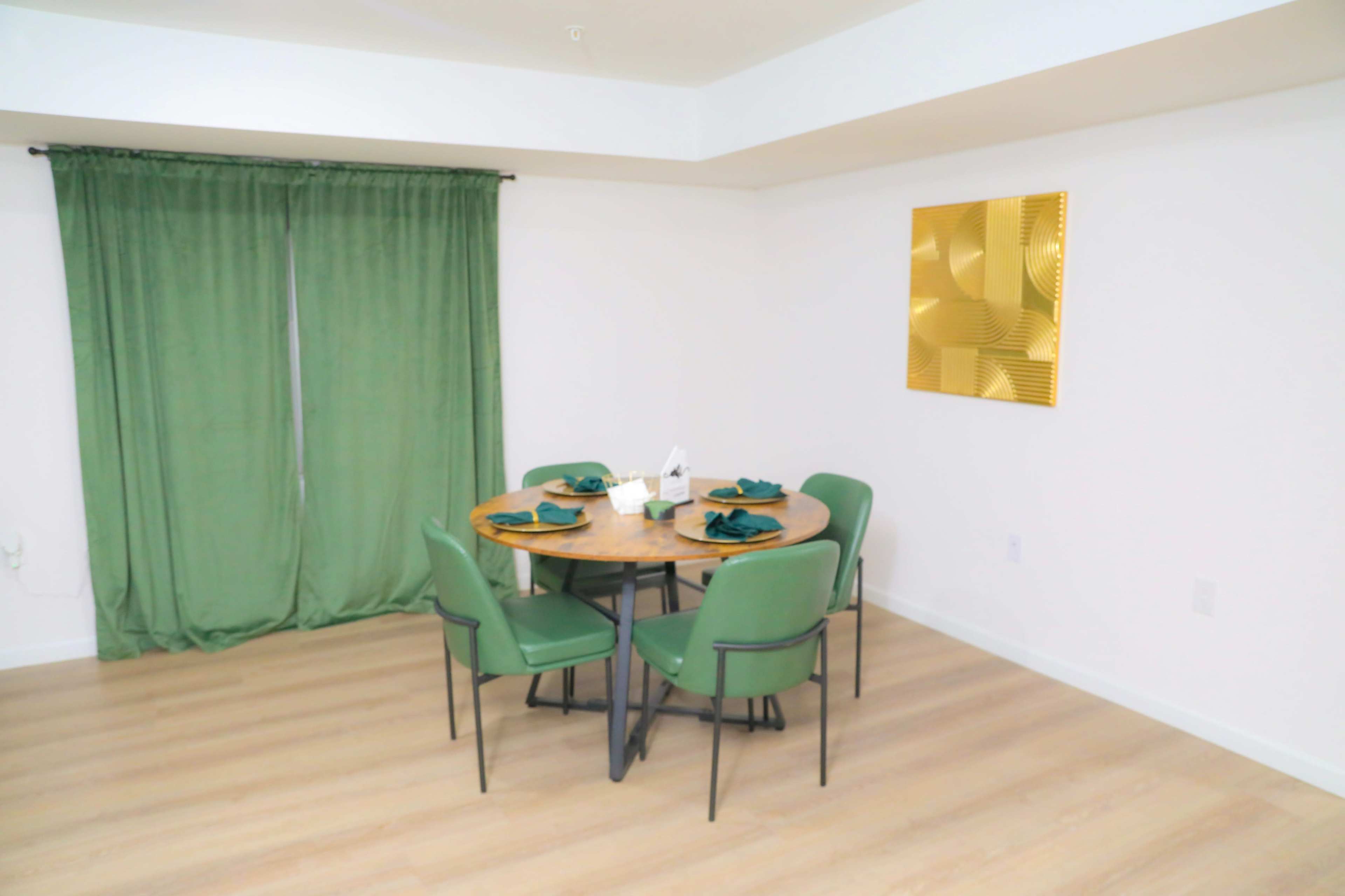 A round dining table with green chairs is set in a brightly lit room, accompanied by curtains and a wall art piece.