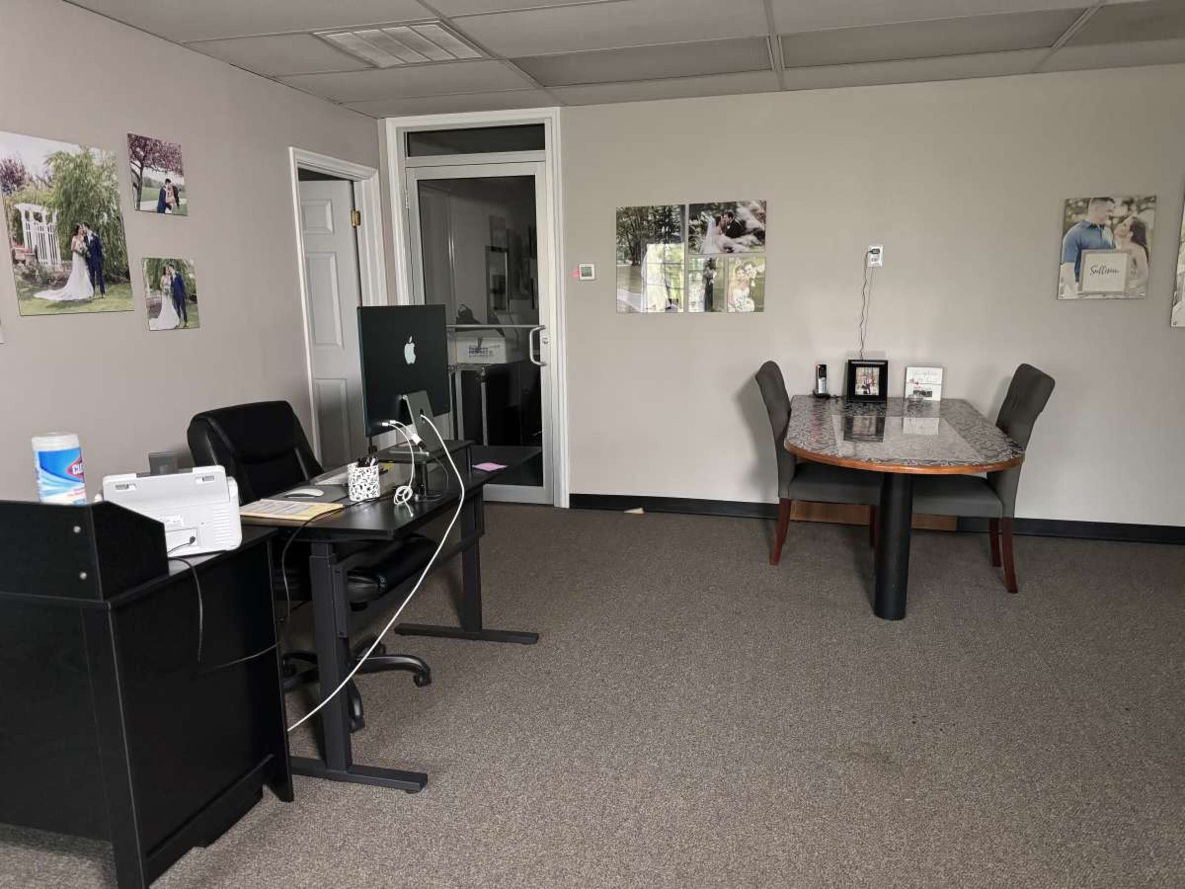 The image shows an office space featuring a desk with a computer, a table with chairs, and several framed photographs on the walls.