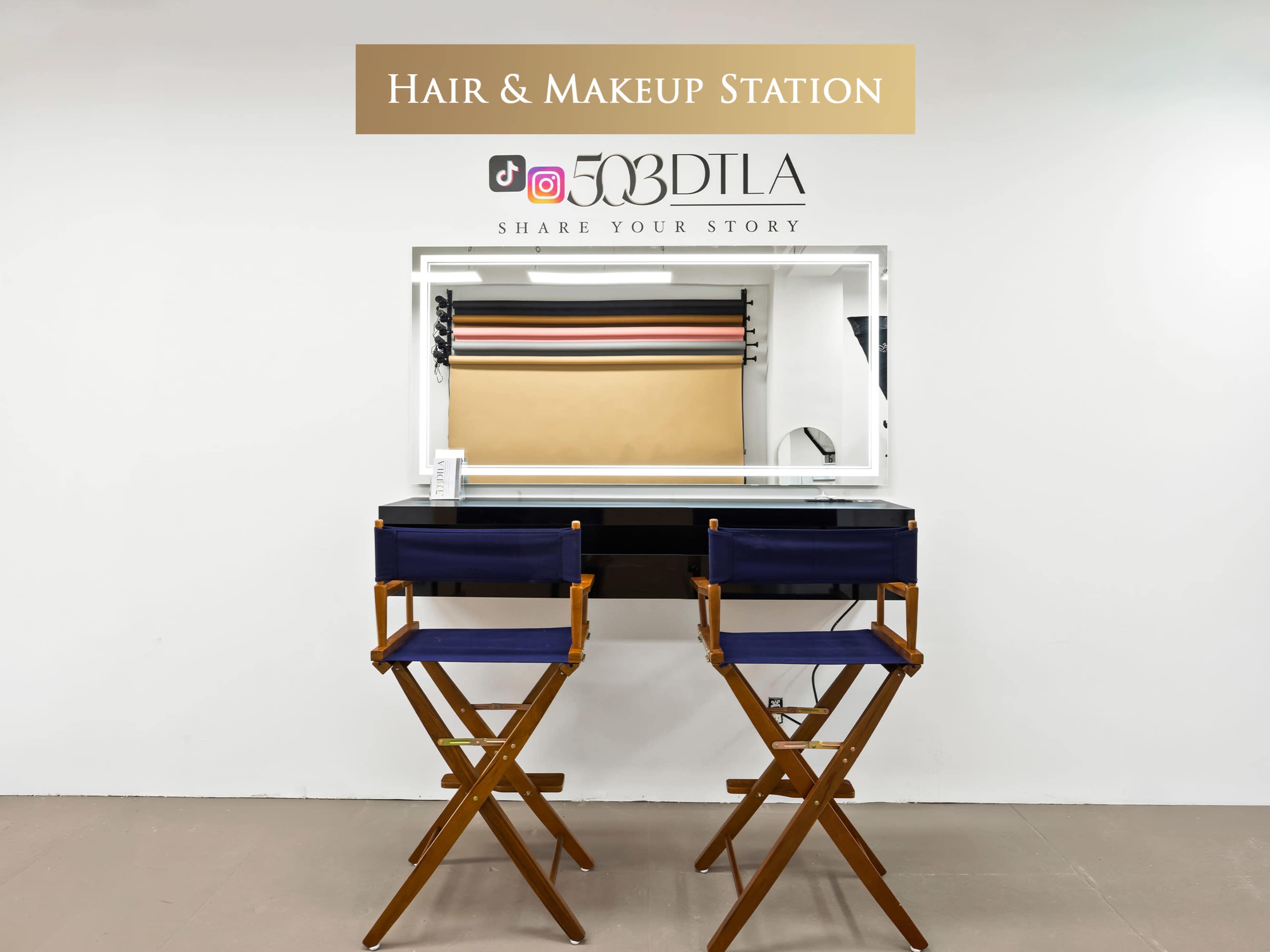 The image shows a hair and makeup station with two director's chairs facing a mirror, framed by colorful backdrop rolls.