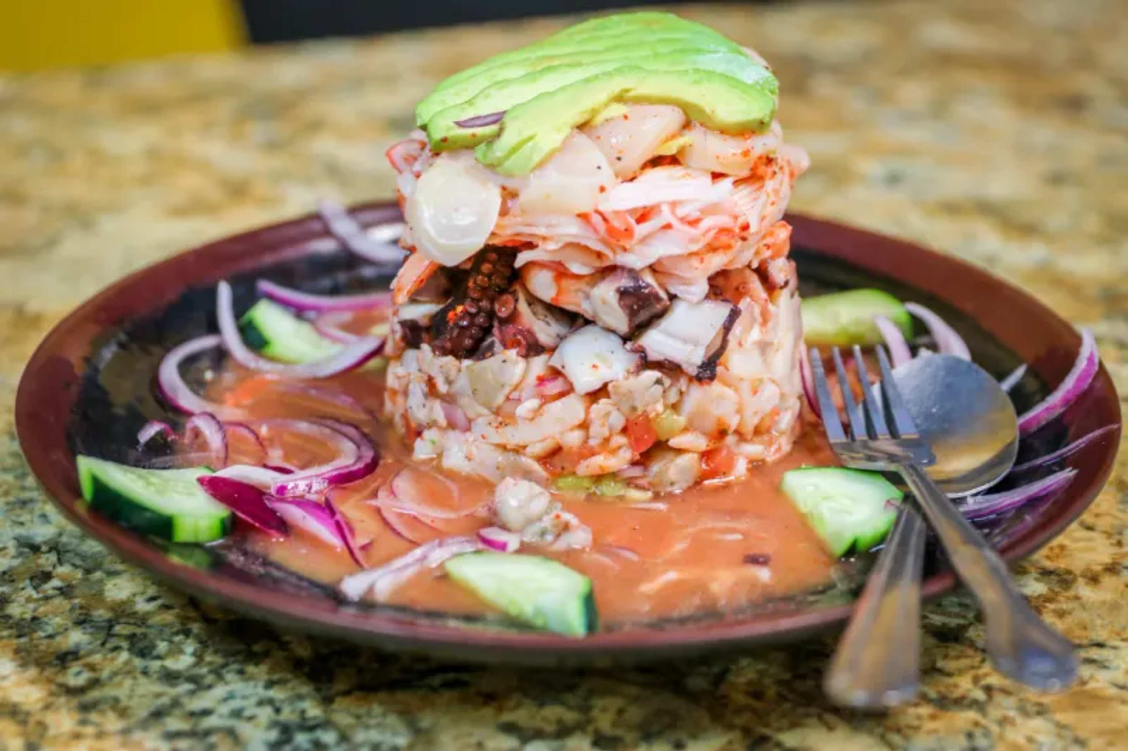 A layered seafood dish featuring octopus, shrimp, and vegetables, garnished with avocado and served on a plate with cucumbers and red onion.