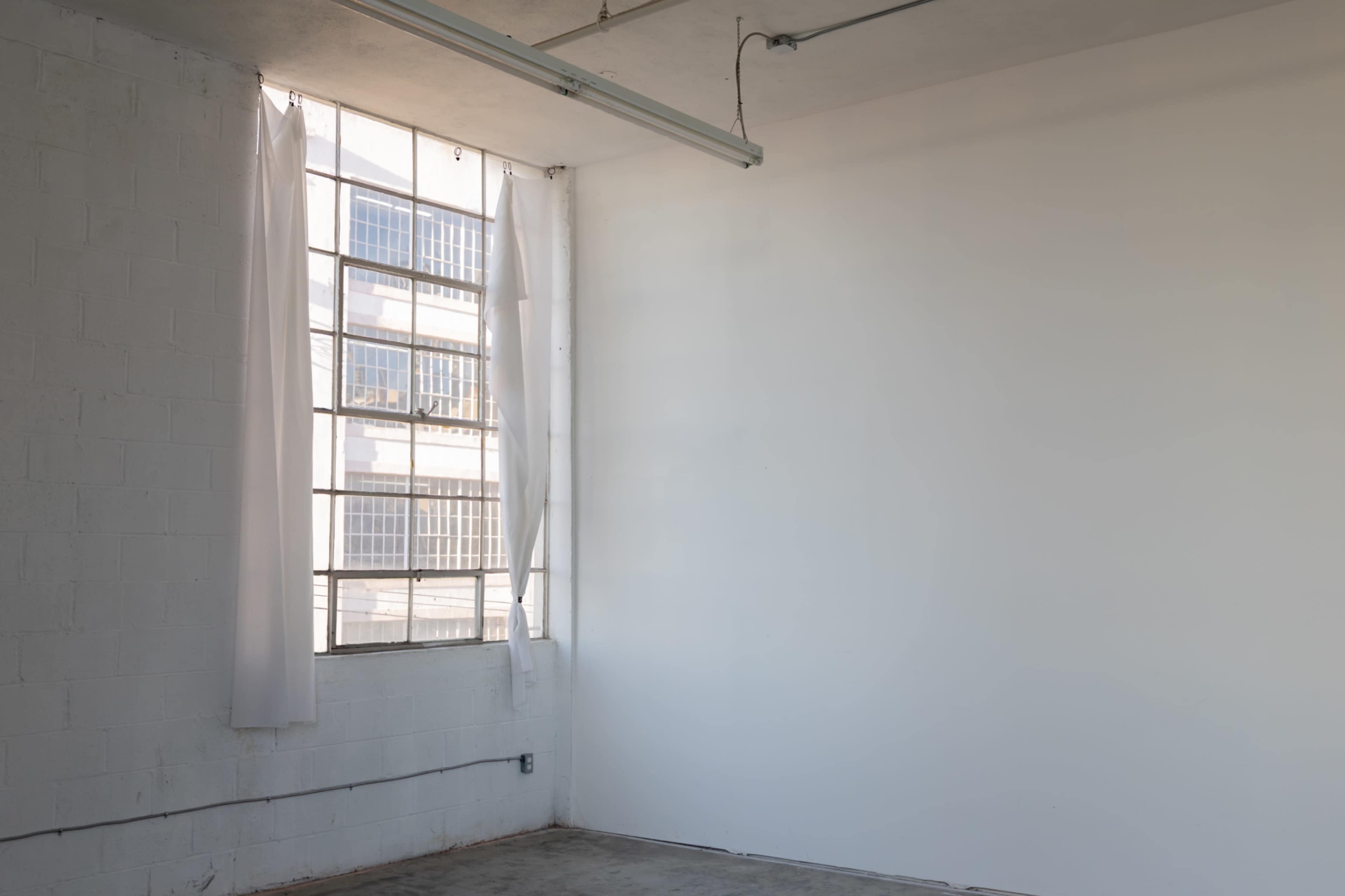 A large window with sheer curtains is set in a bare, white-walled room with a concrete floor.