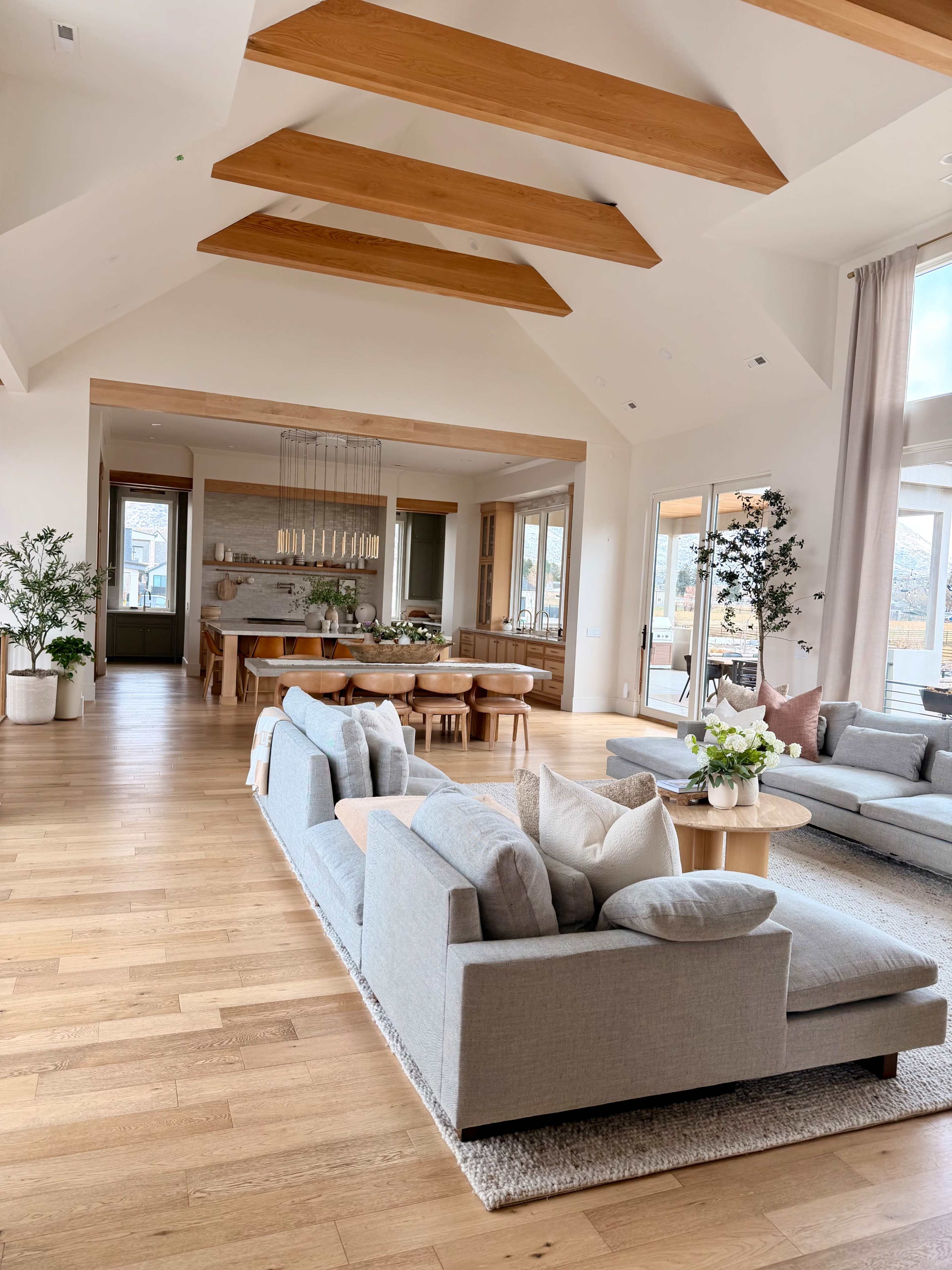 The image features a spacious living area with high ceilings, wooden beams, a large sectional sofa, and a dining table set for multiple people.