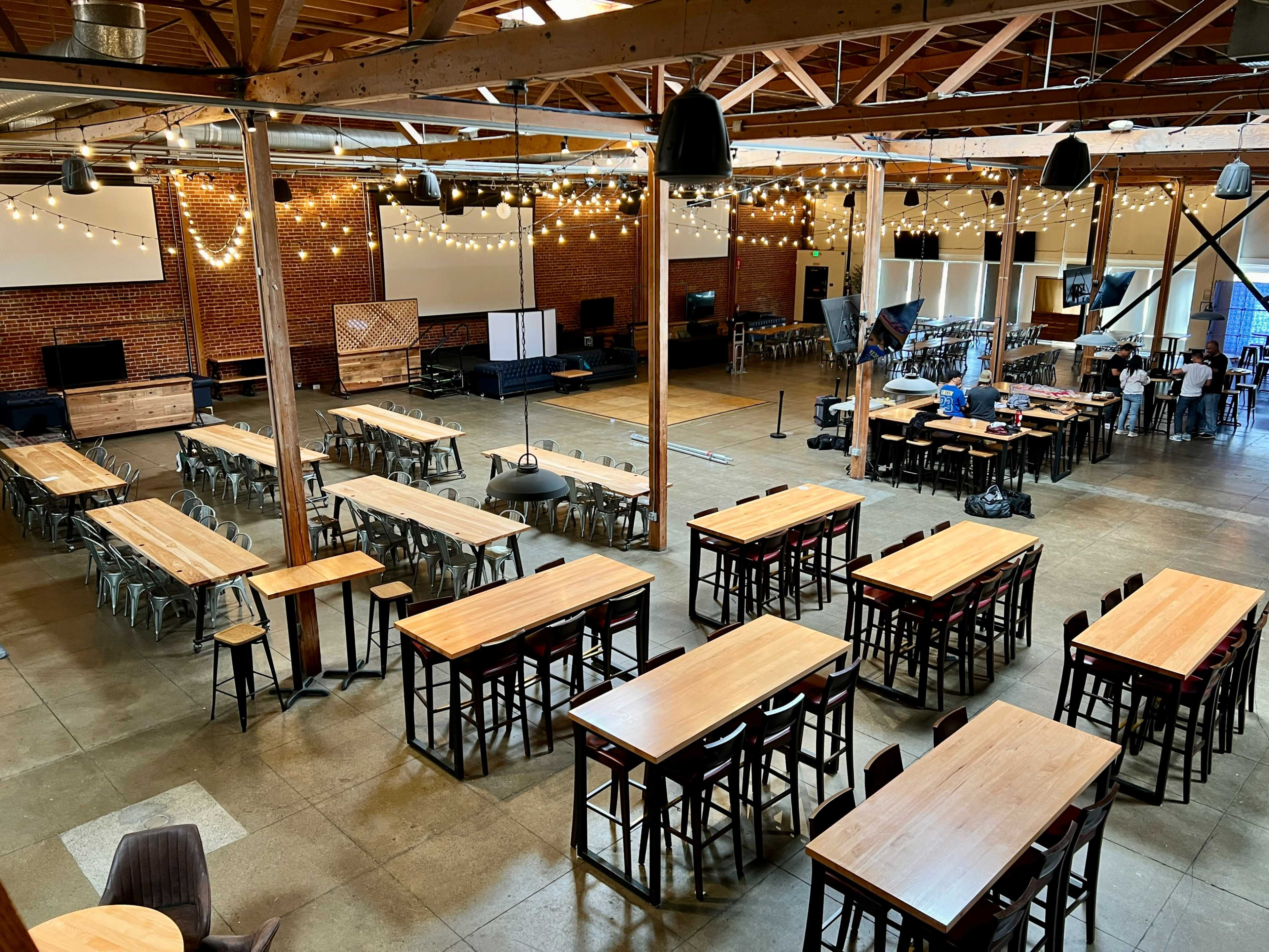 The image shows a spacious event venue with long wooden tables arranged in rows, string lights overhead, brick walls, and a stage area at one end.