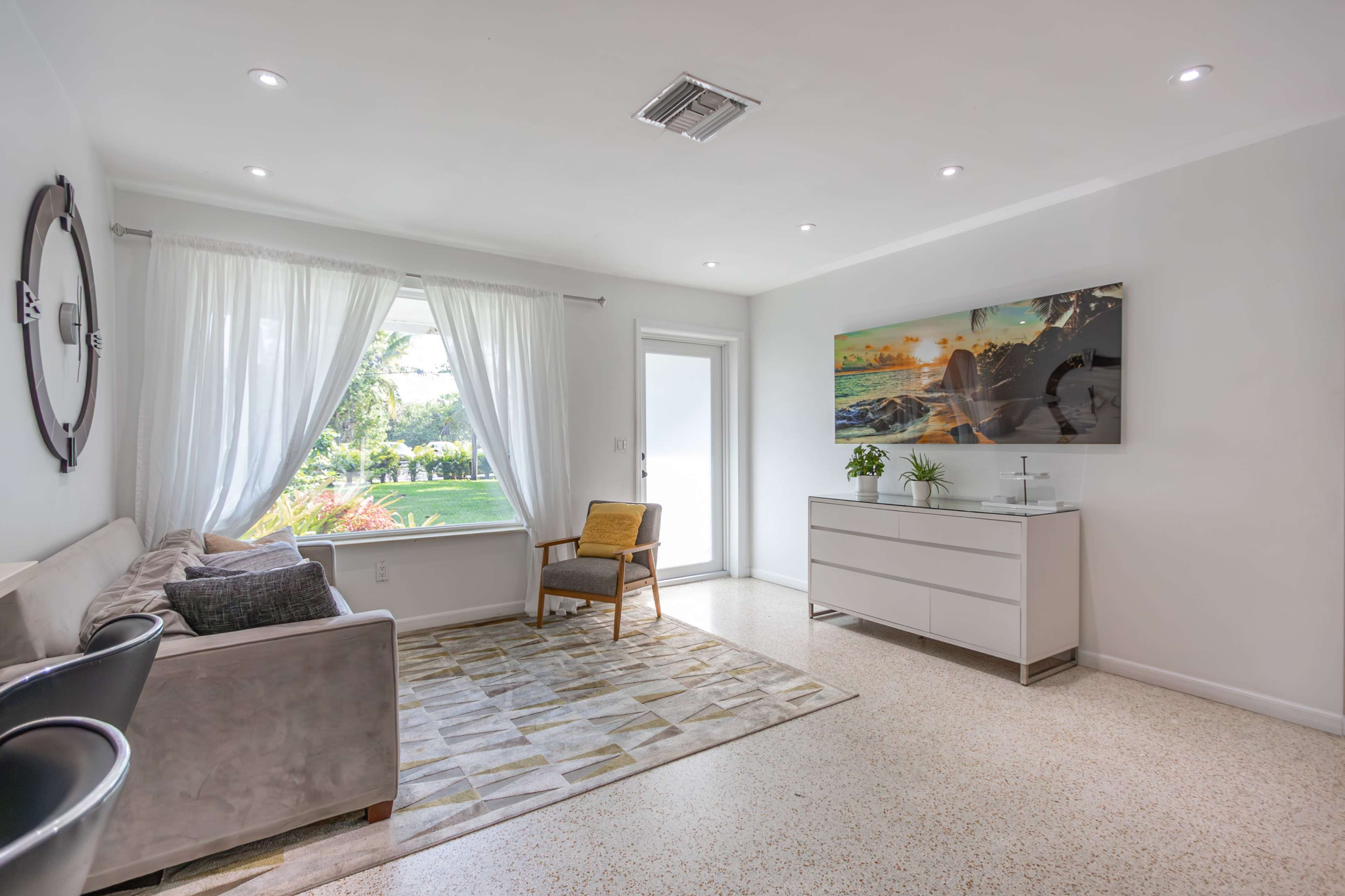 The scene depicts a modern living room with a sofa, an armchair, a white media console, and a large window overlooking a garden.