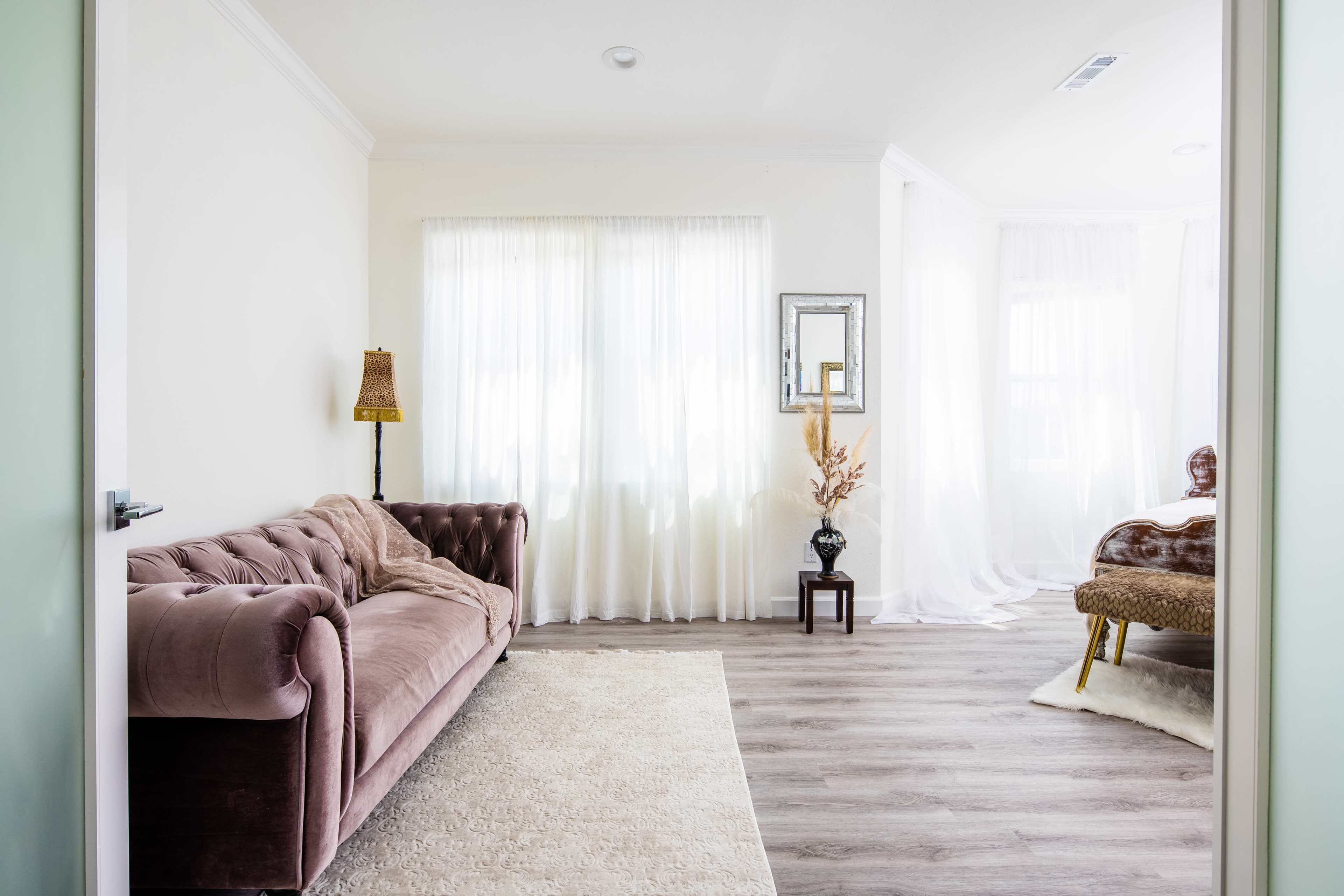 A well-lit room features a velvet sofa, a decorative mirror, and a bedside table with a vase, complemented by sheer curtains and wooden flooring.