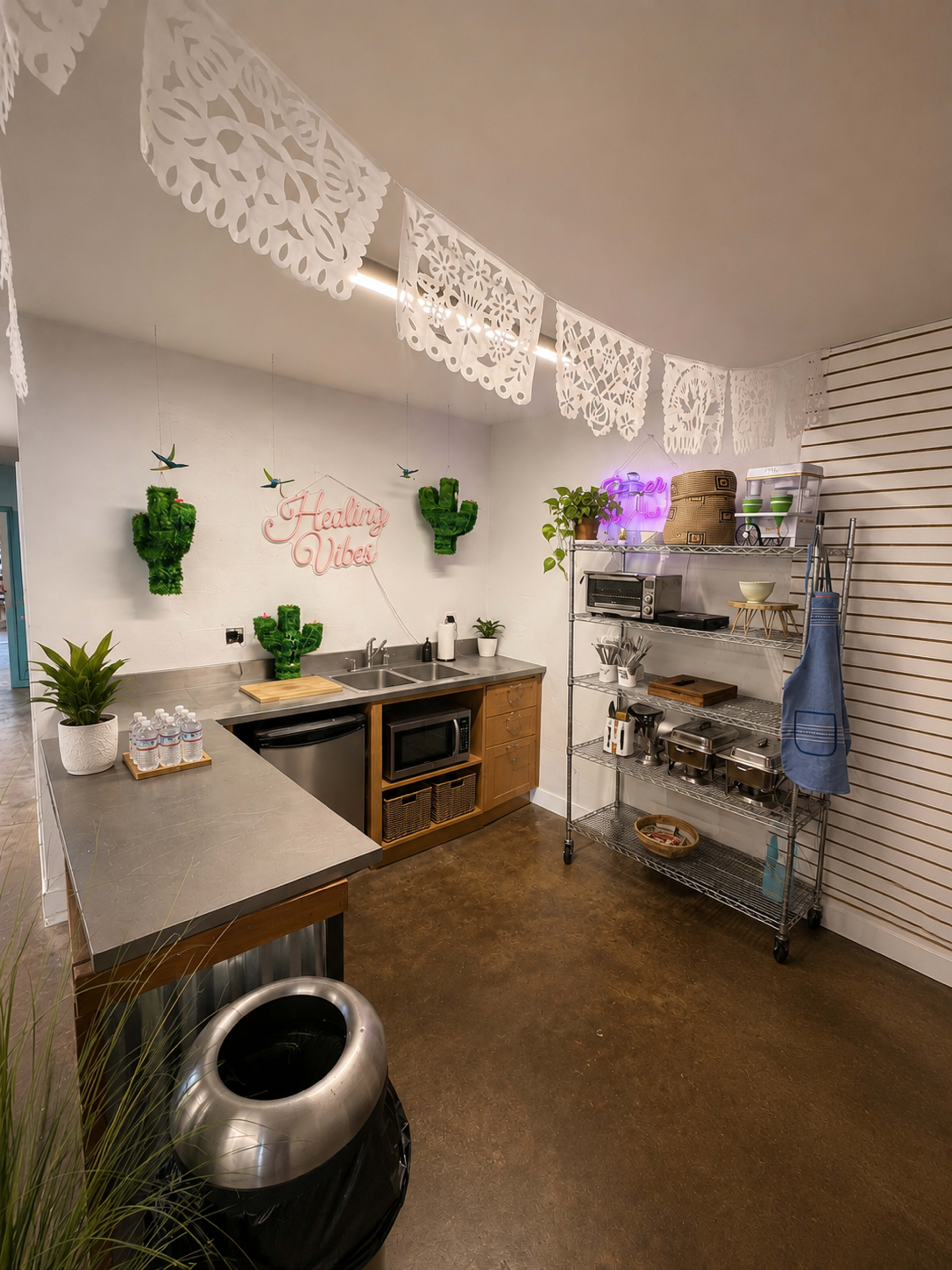 The image shows a cozy kitchen space featuring a stainless steel countertop, a microwave, plants, and decorative paper banners.