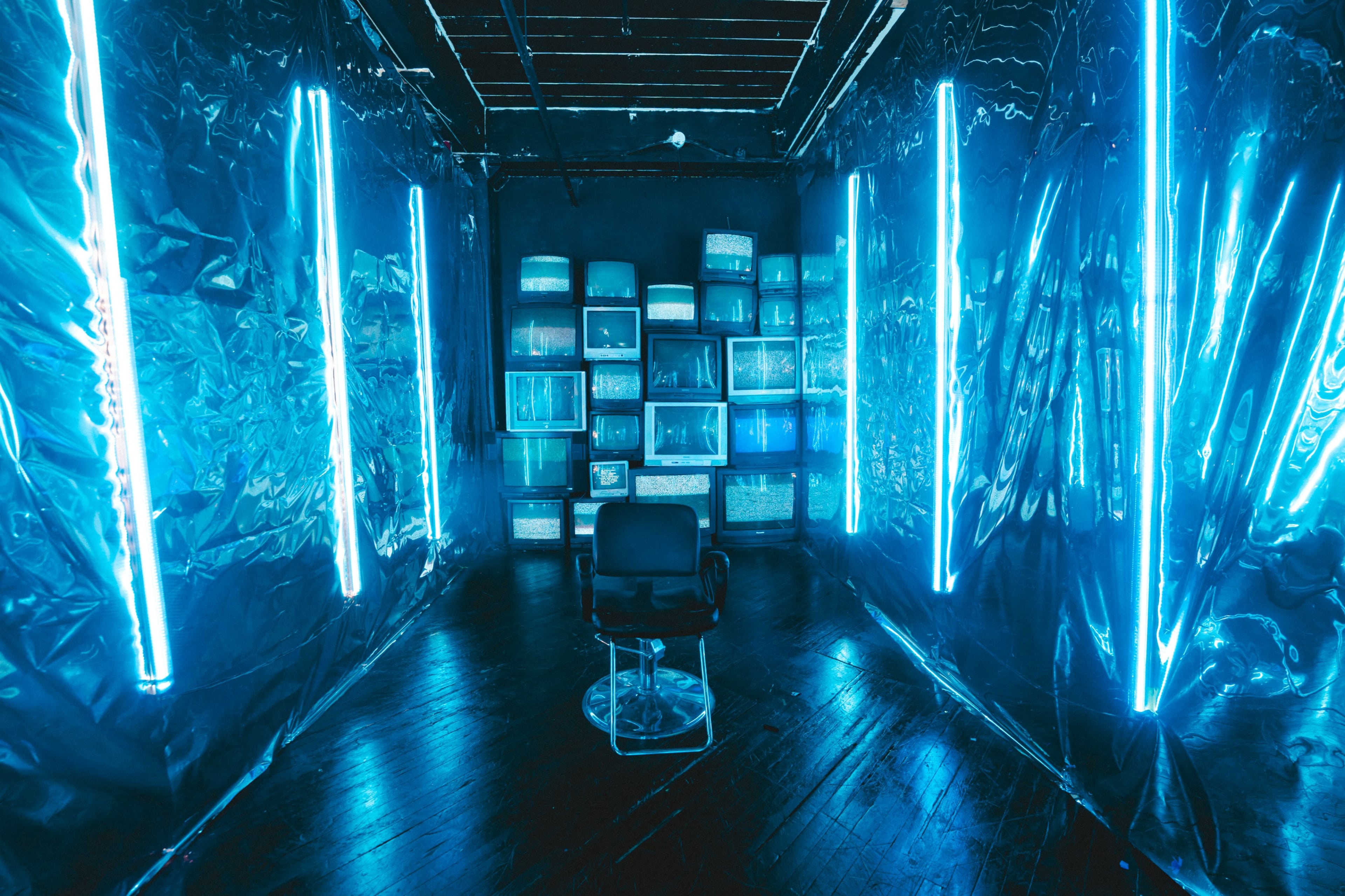 The image shows a dimly lit room with blue neon lights, featuring a circular chair in the center and a wall of stacked old television screens.