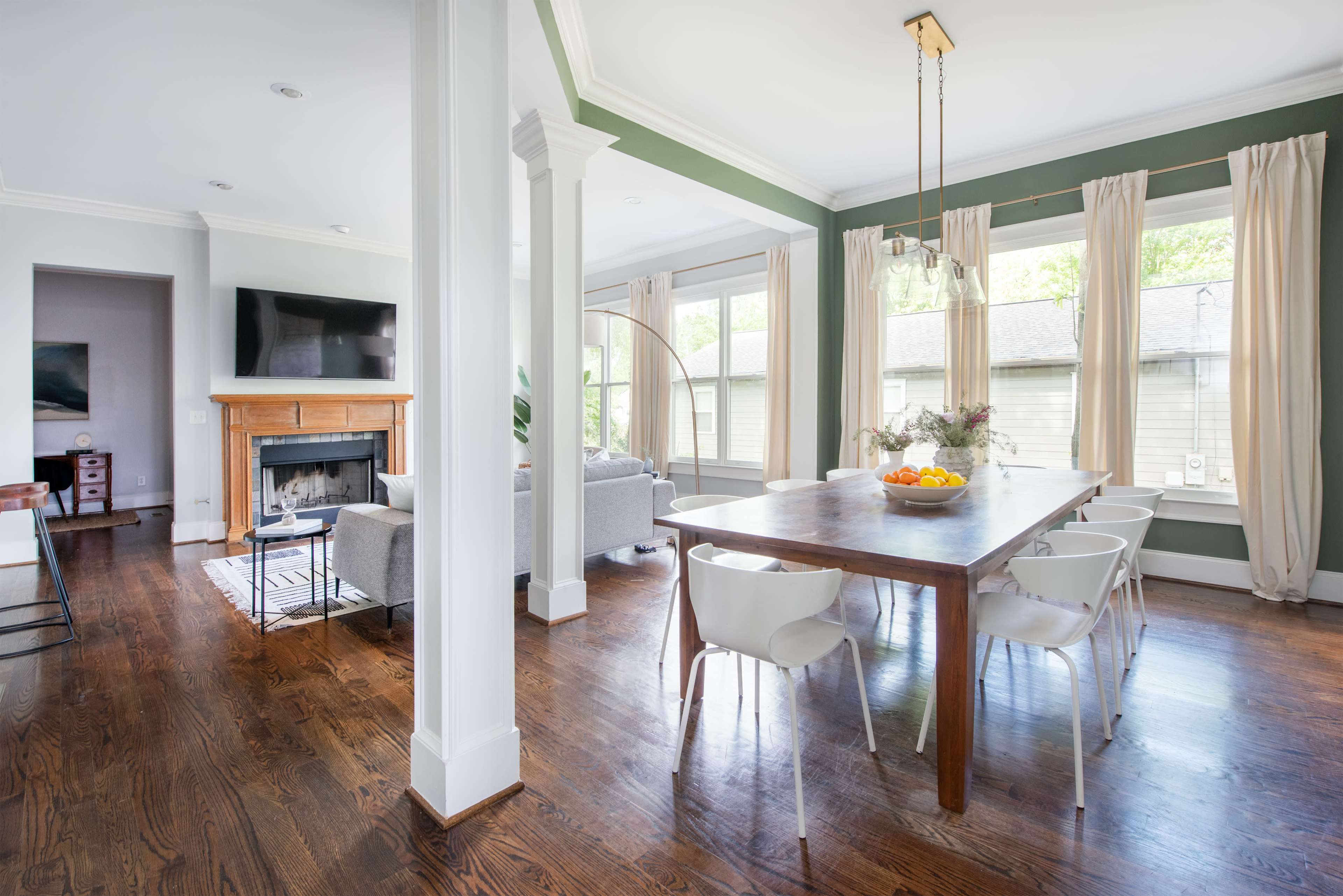 A dining area features a wooden table surrounded by white chairs, with a fireplace and large windows in a bright, open layout.