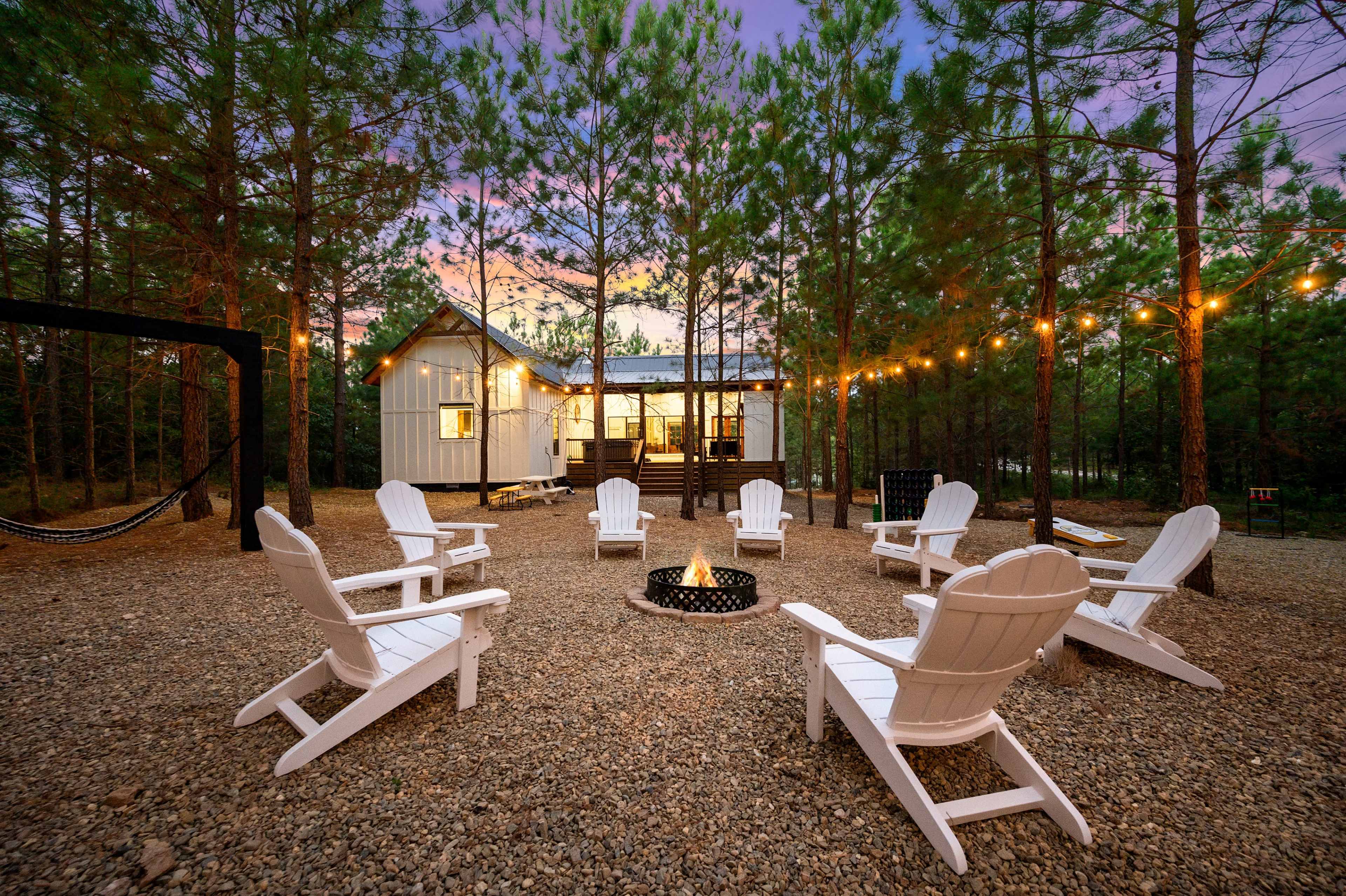 A gravel area surrounded by trees, featuring a fire pit and several white Adirondack chairs arranged in a circle, with a cabin illuminated in the background.