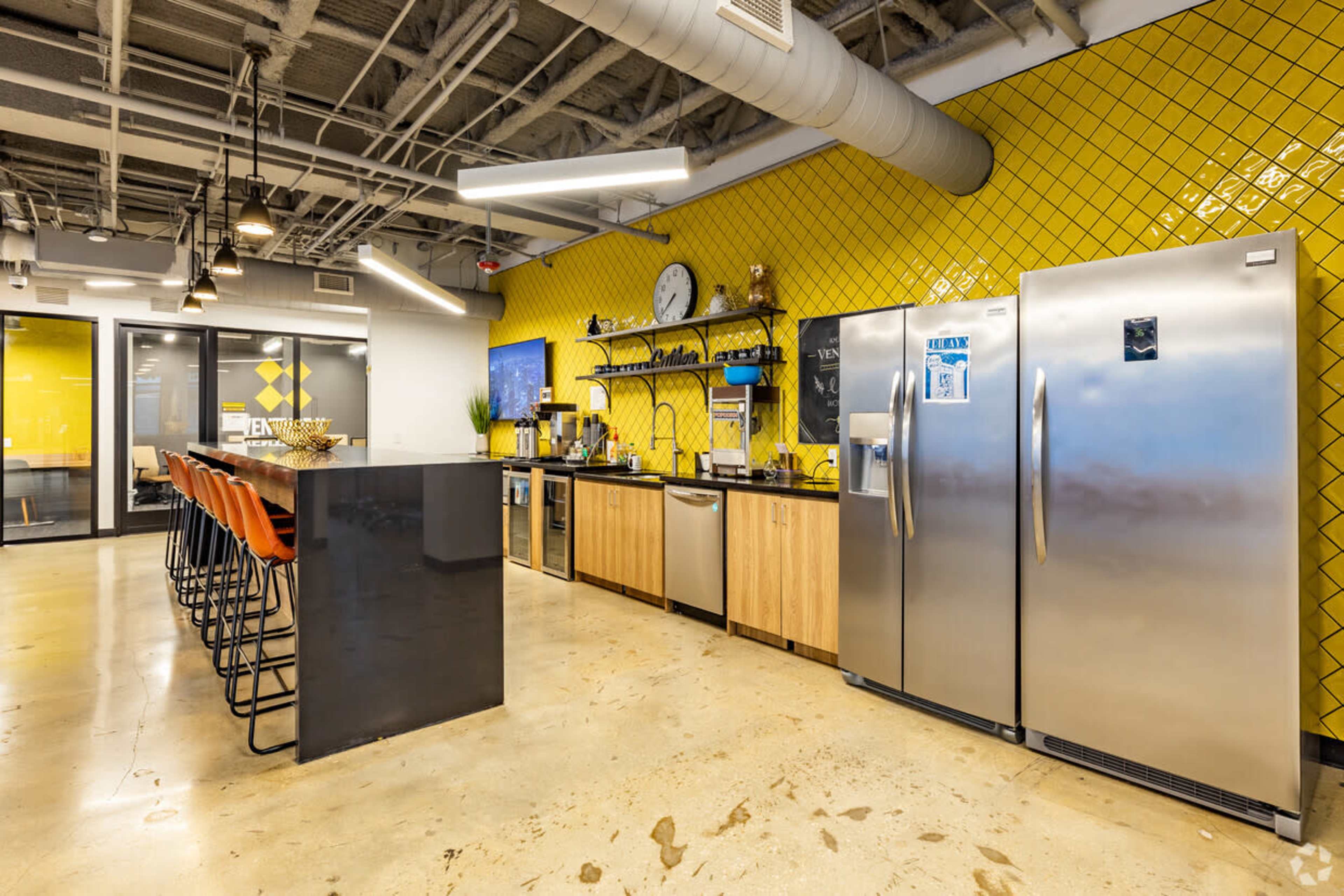 A modern kitchen area with yellow tiled walls, stainless steel refrigerators, and a long bar with high stools.