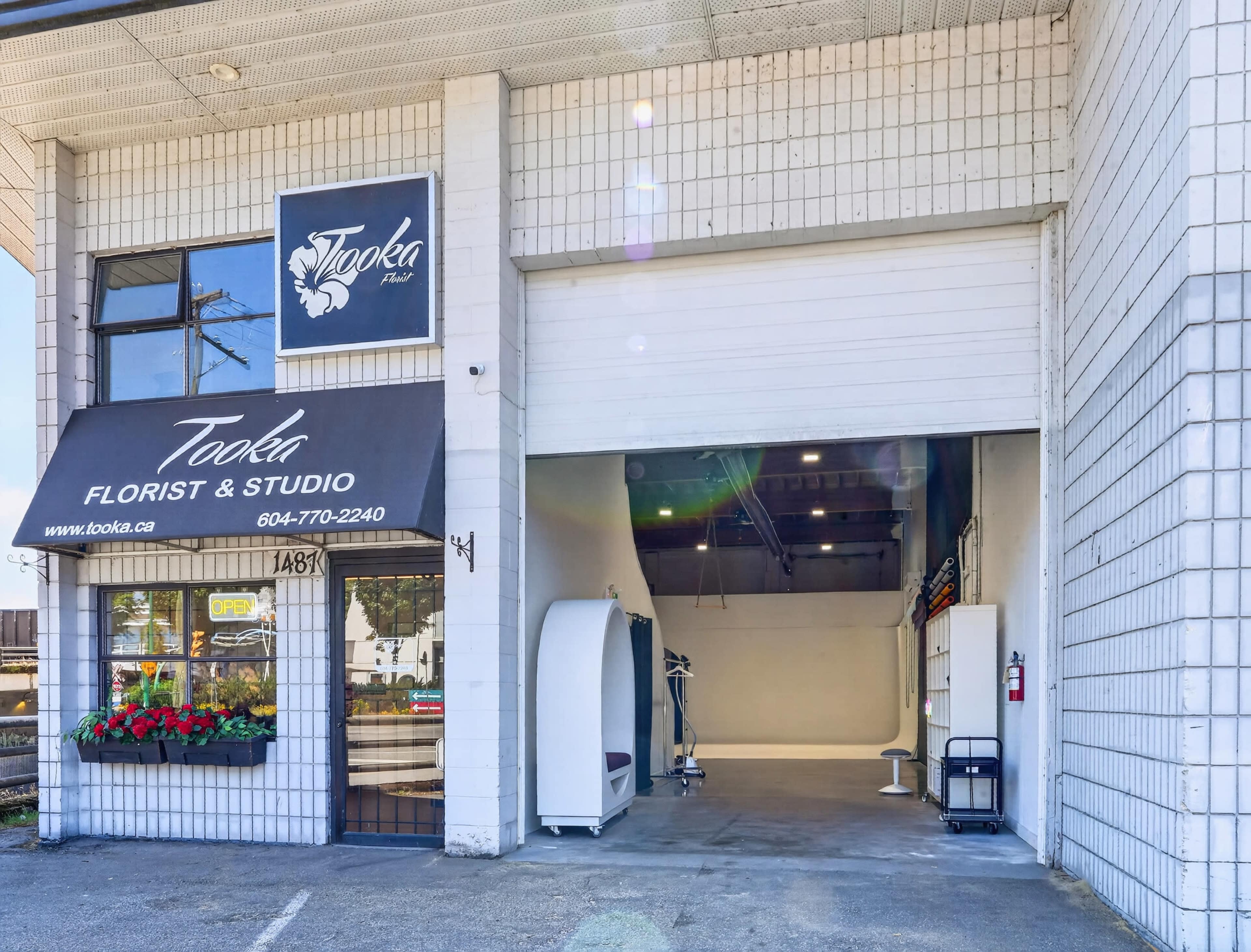 The image shows the entrance of a florist and studio named "Tooka," featuring a large opening, a black awning with the business name, and potted flowers.