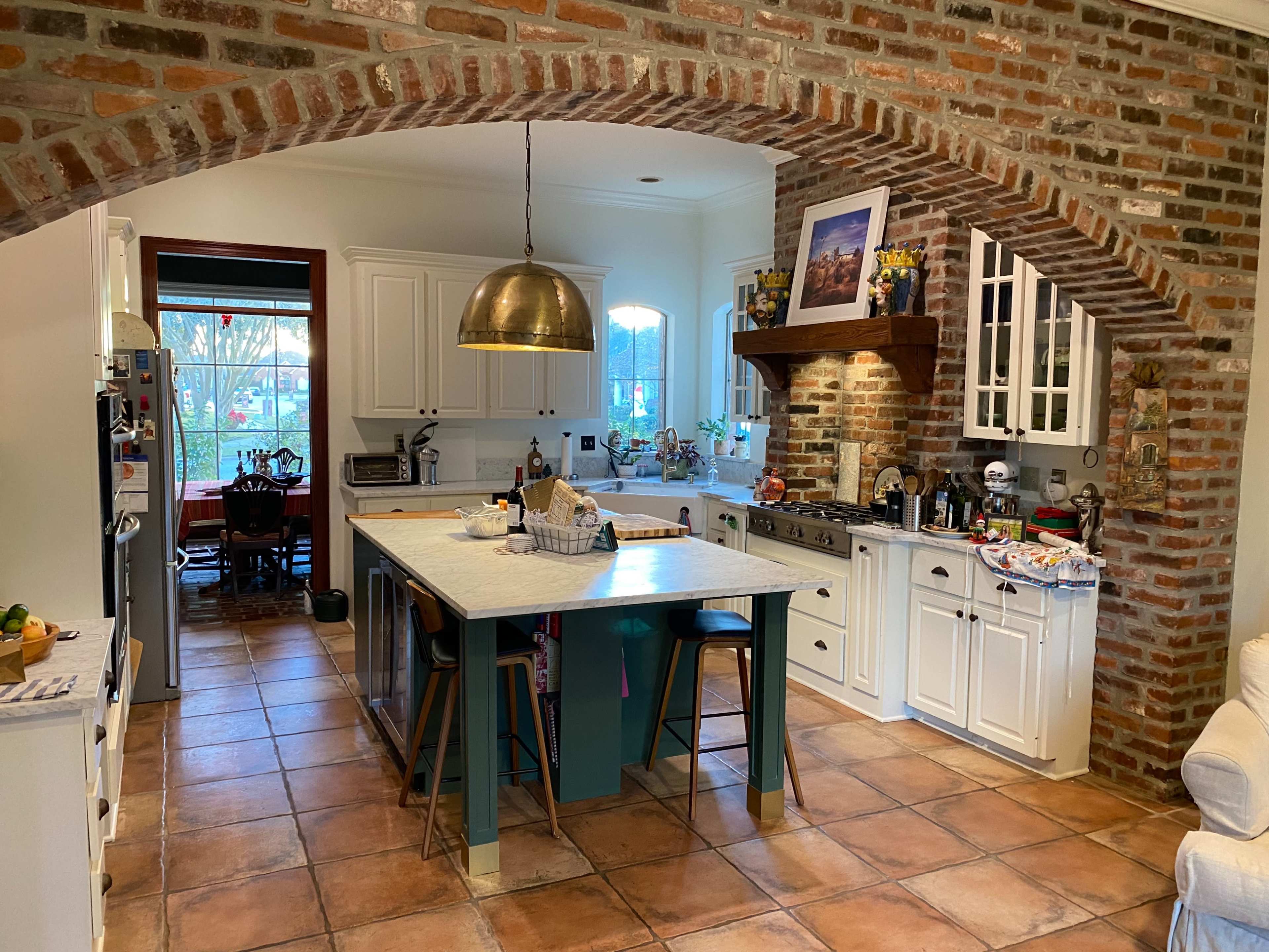 The kitchen features an arched brick entryway, a large island with bar stools, and white cabinetry with a mixture of modern appliances and rustic decor.