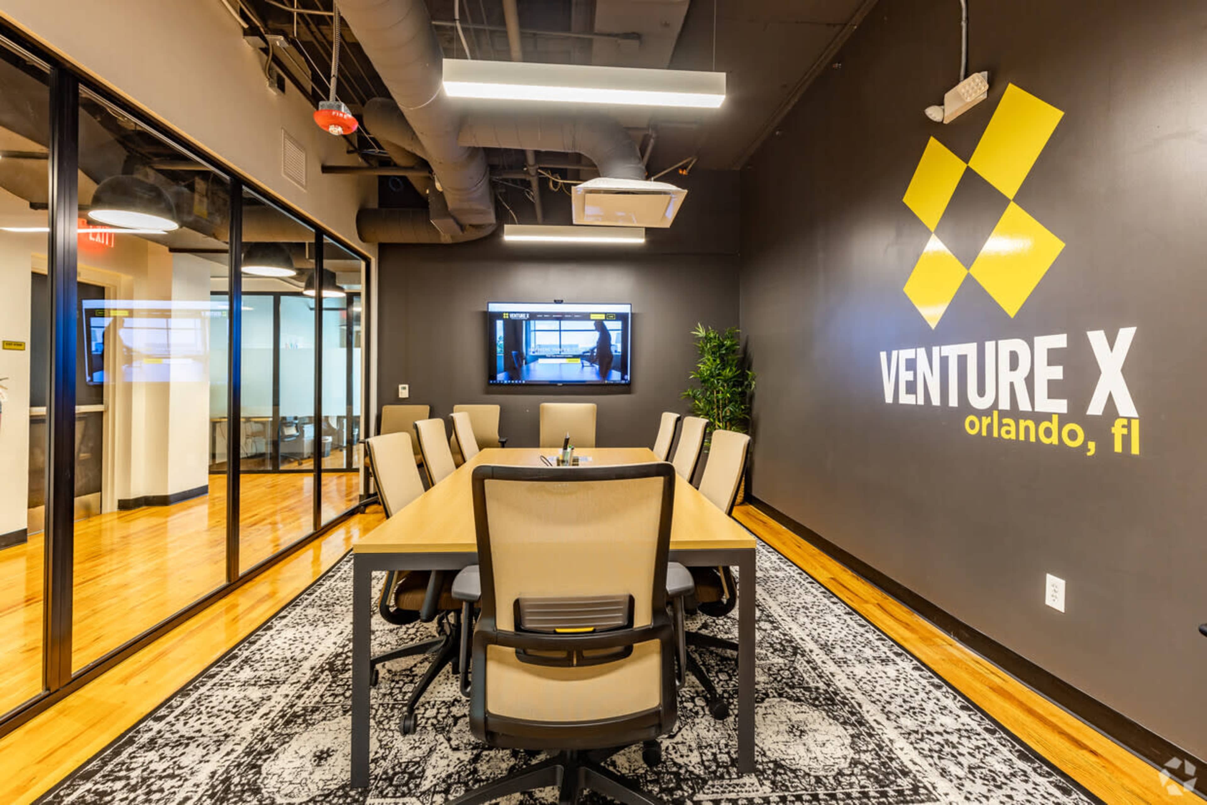 A modern conference room with a large wooden table, ergonomic chairs, and a television screen on the wall, featuring the "Venture X" logo prominently displayed.
