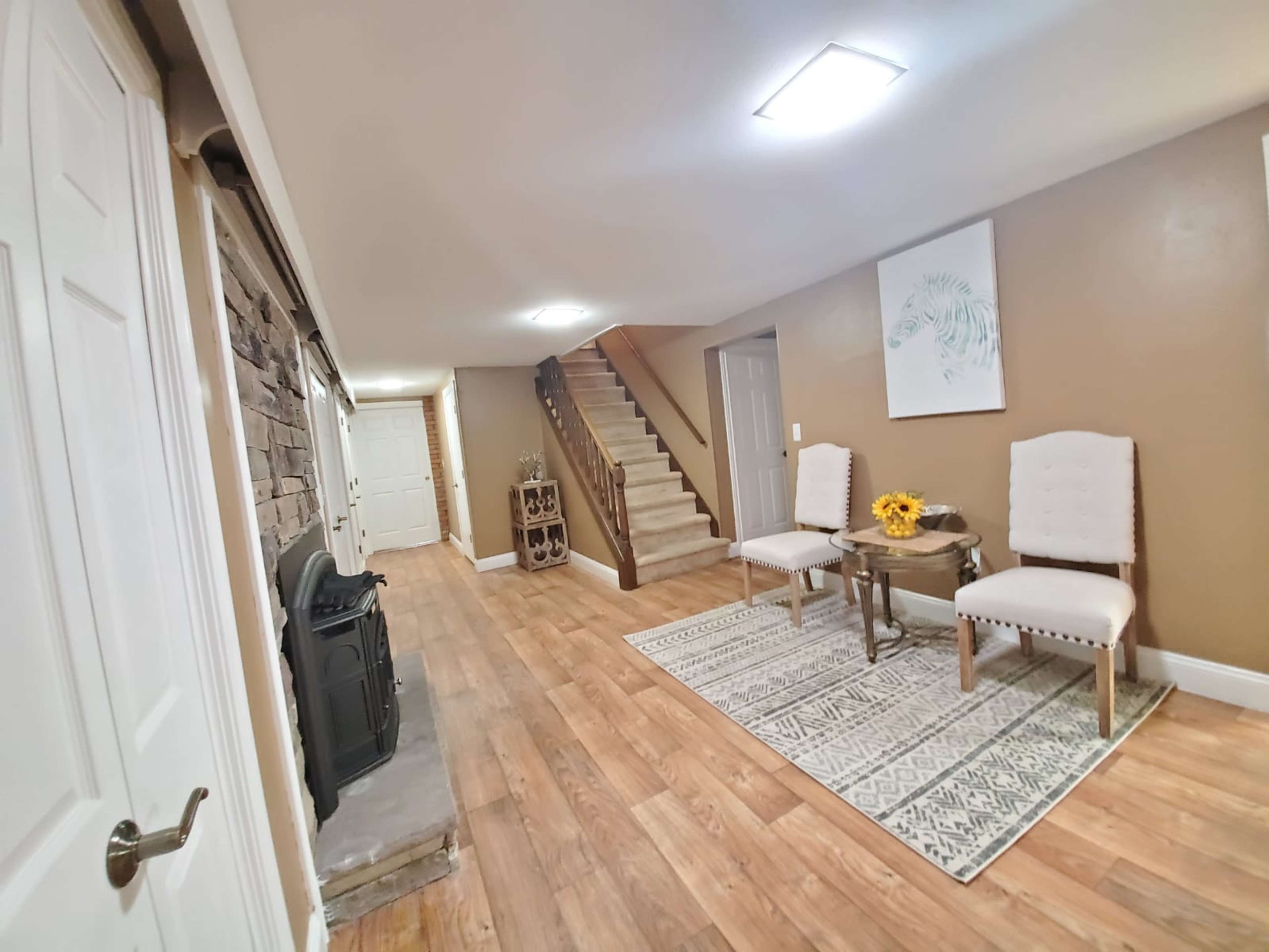 The image shows a well-lit basement area with a staircase, two chairs, a small table with sunflowers, and a decorative rug.