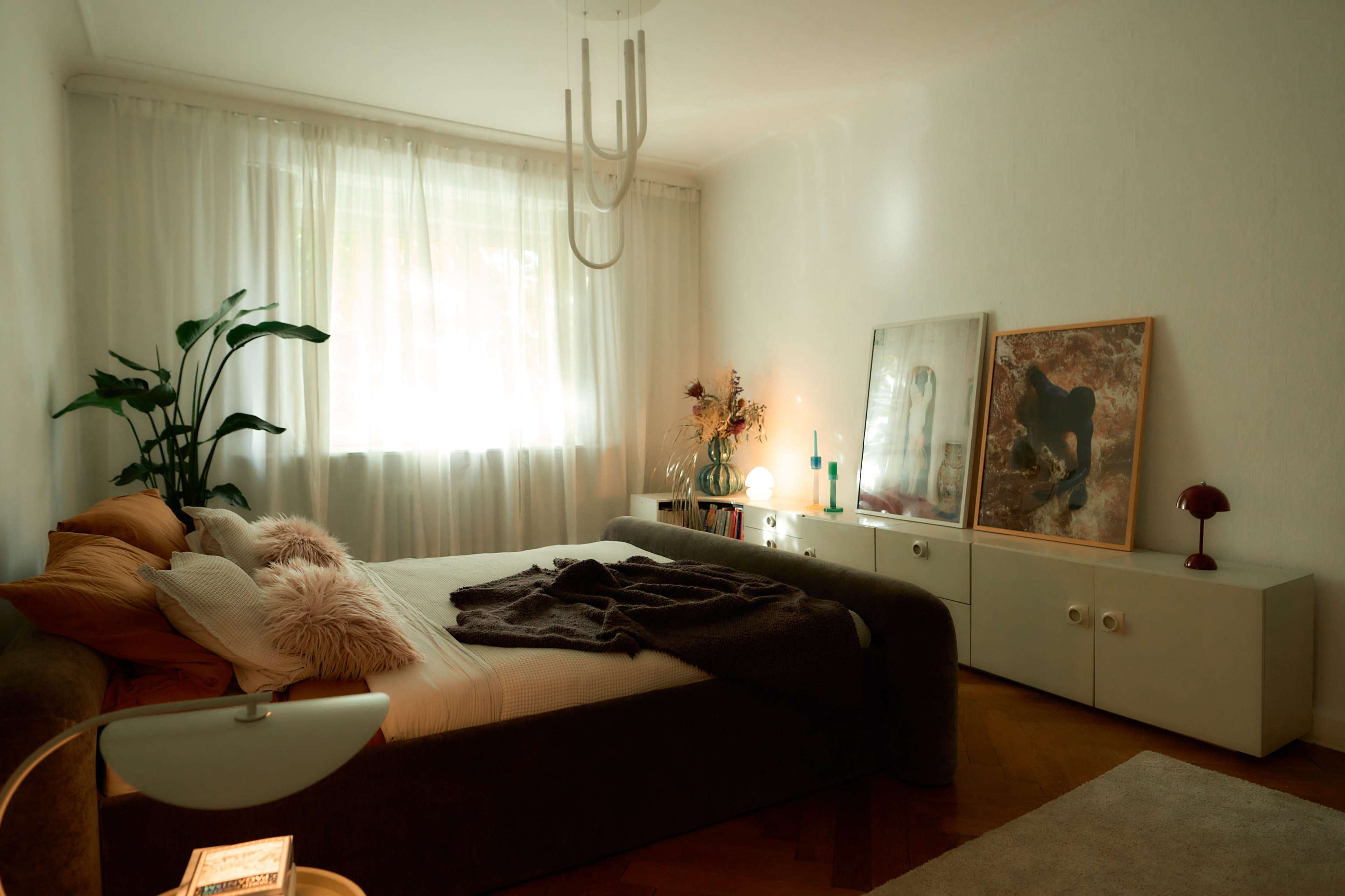 A softly lit bedroom features a bed with decorative pillows, a nearby plant, and two framed artworks displayed on a low dresser.