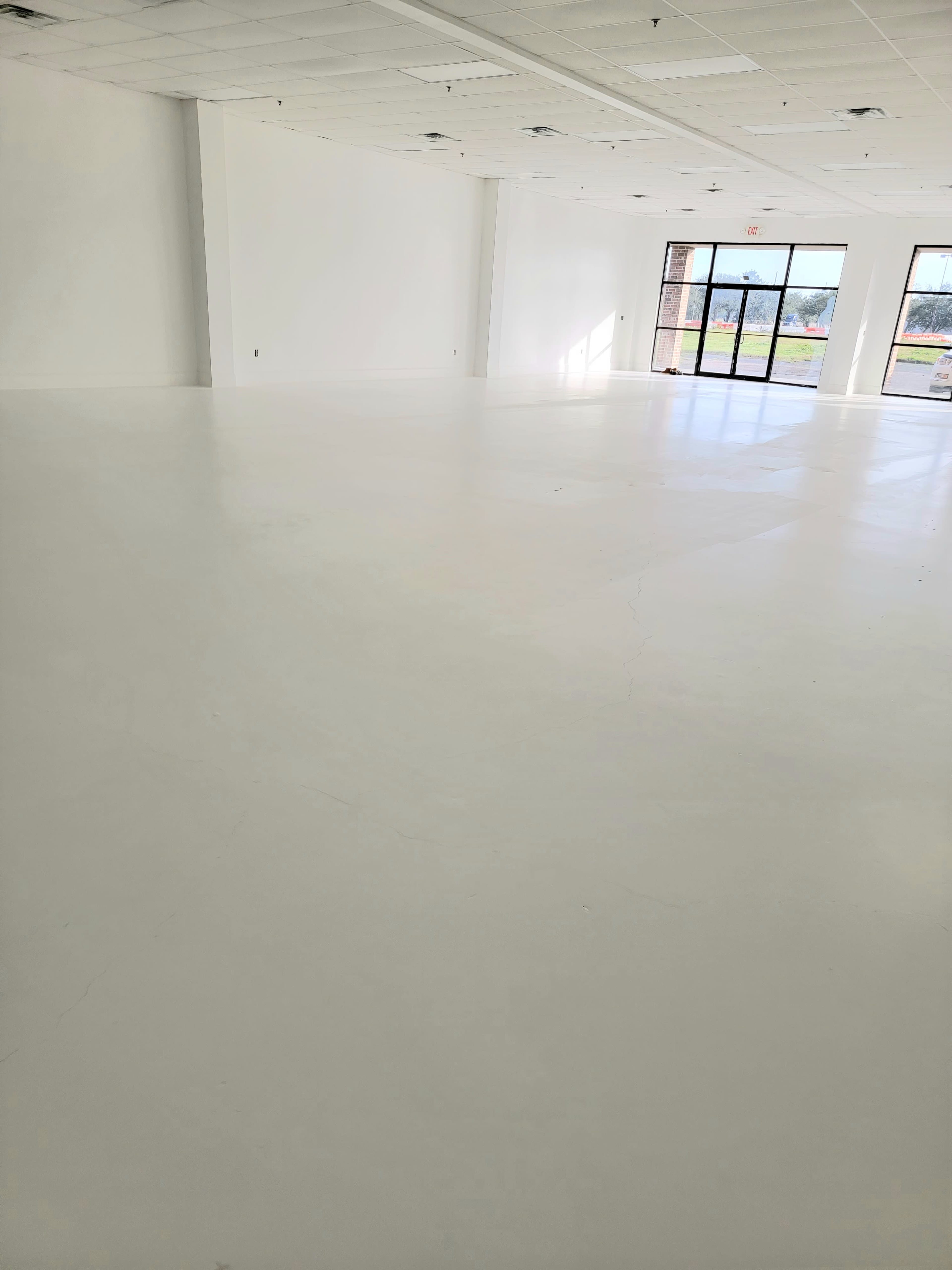 The image shows an empty room with white walls and a smooth white floor, featuring large windows that allow natural light to enter.