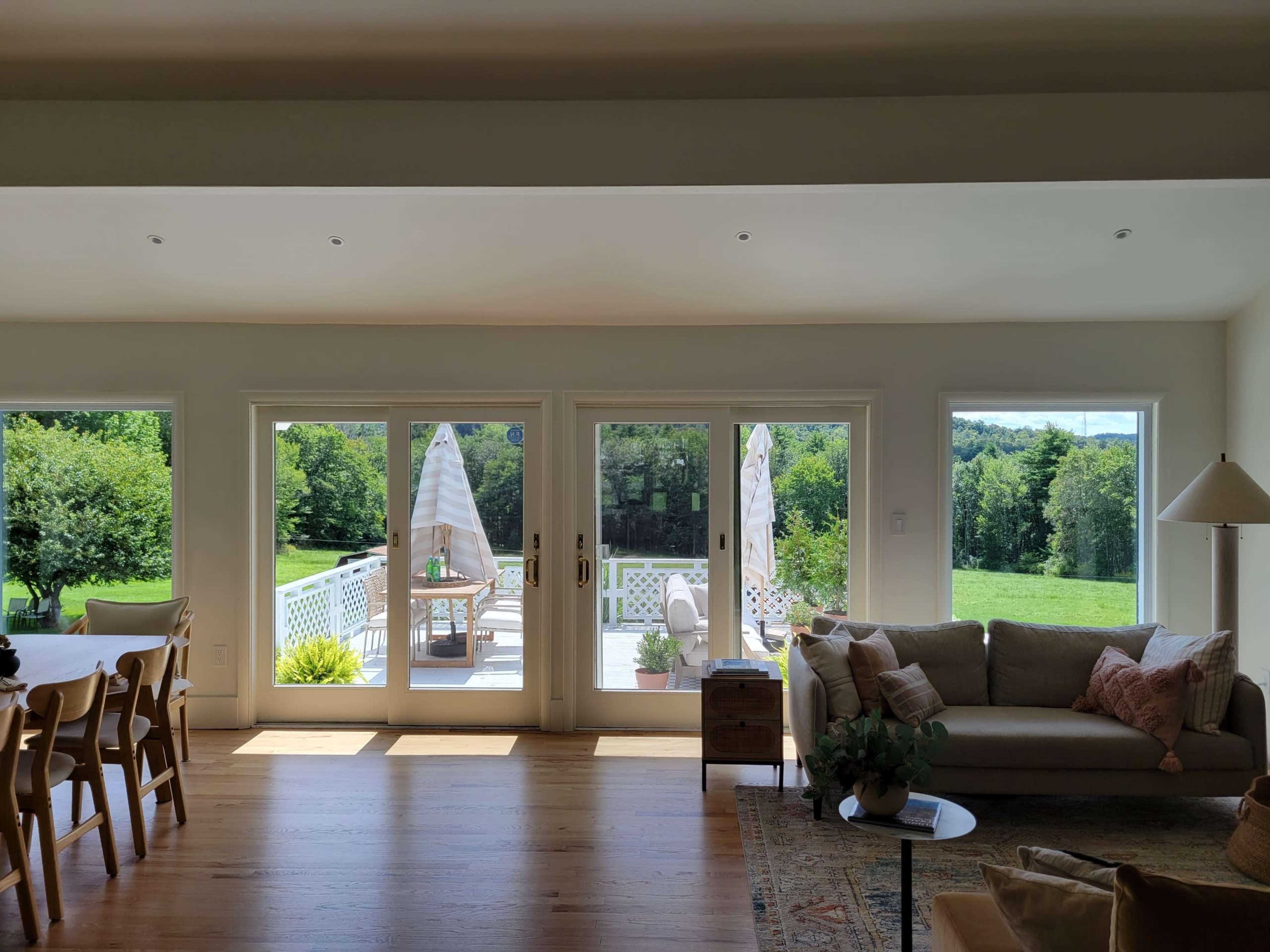 A bright living room features large windows leading to a deck with outdoor furniture and greenery in the background.